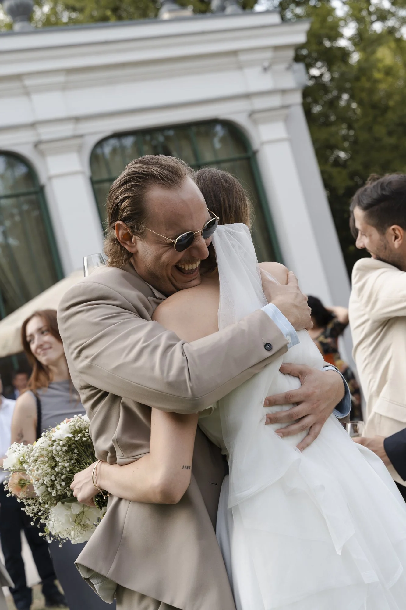 People celebrating outdoors at a wedding, with two individuals hugging and smiling, one holding a bouquet of flowers.