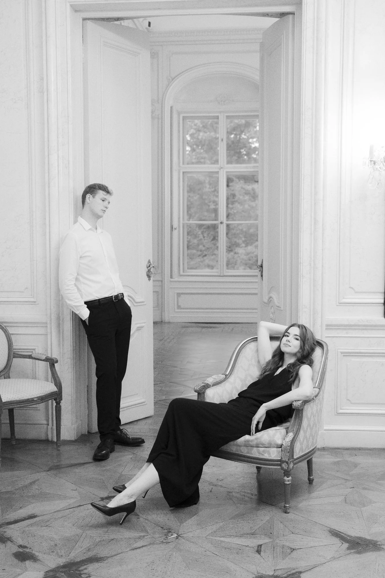 A woman sitting in a vintage armchair with one arm resting on her head and the other on the armrest, wearing a long black dress and high heels, while a man in a white shirt and black pants stands nearby in a grand, well-lit room with large windows and ornate decor.