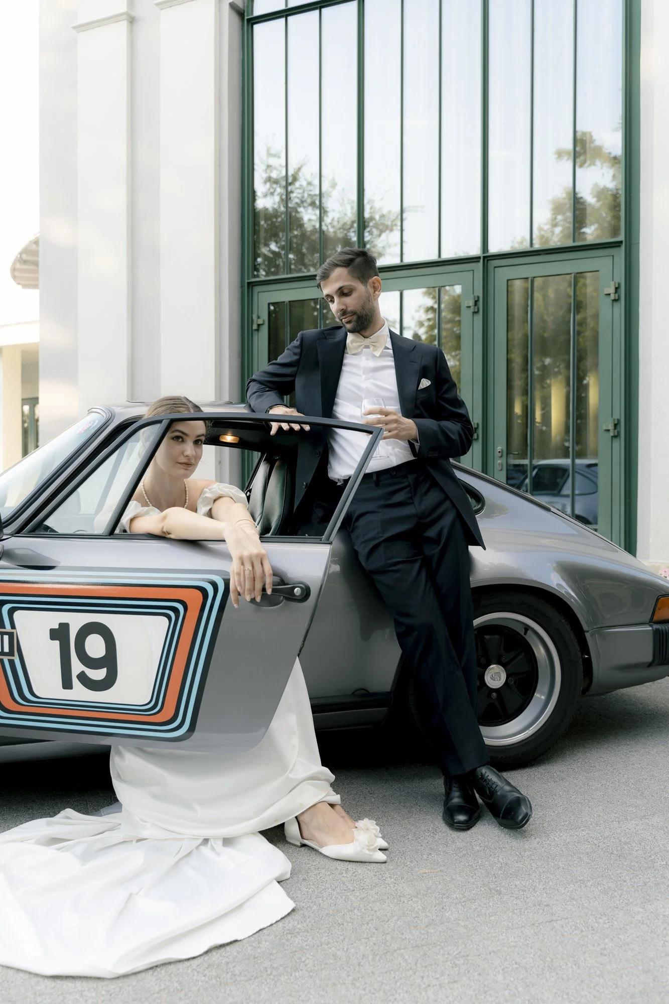 A man in a tuxedo and a woman in a wedding dress leaning on a vintage Porsche car with a racing number, outside a modern building with large glass windows.