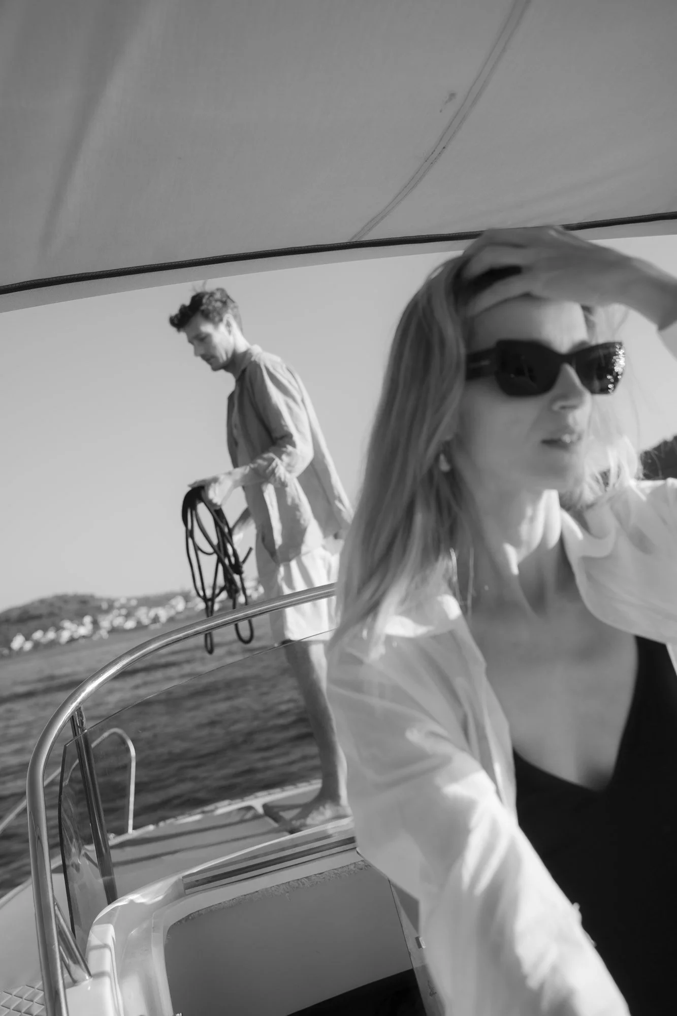 A black-and-white photo of a woman with sunglasses and a man on a boat. The woman is in the foreground, and the man is in the background holding a coiled rope, with a landscape of water and distant land behind them.