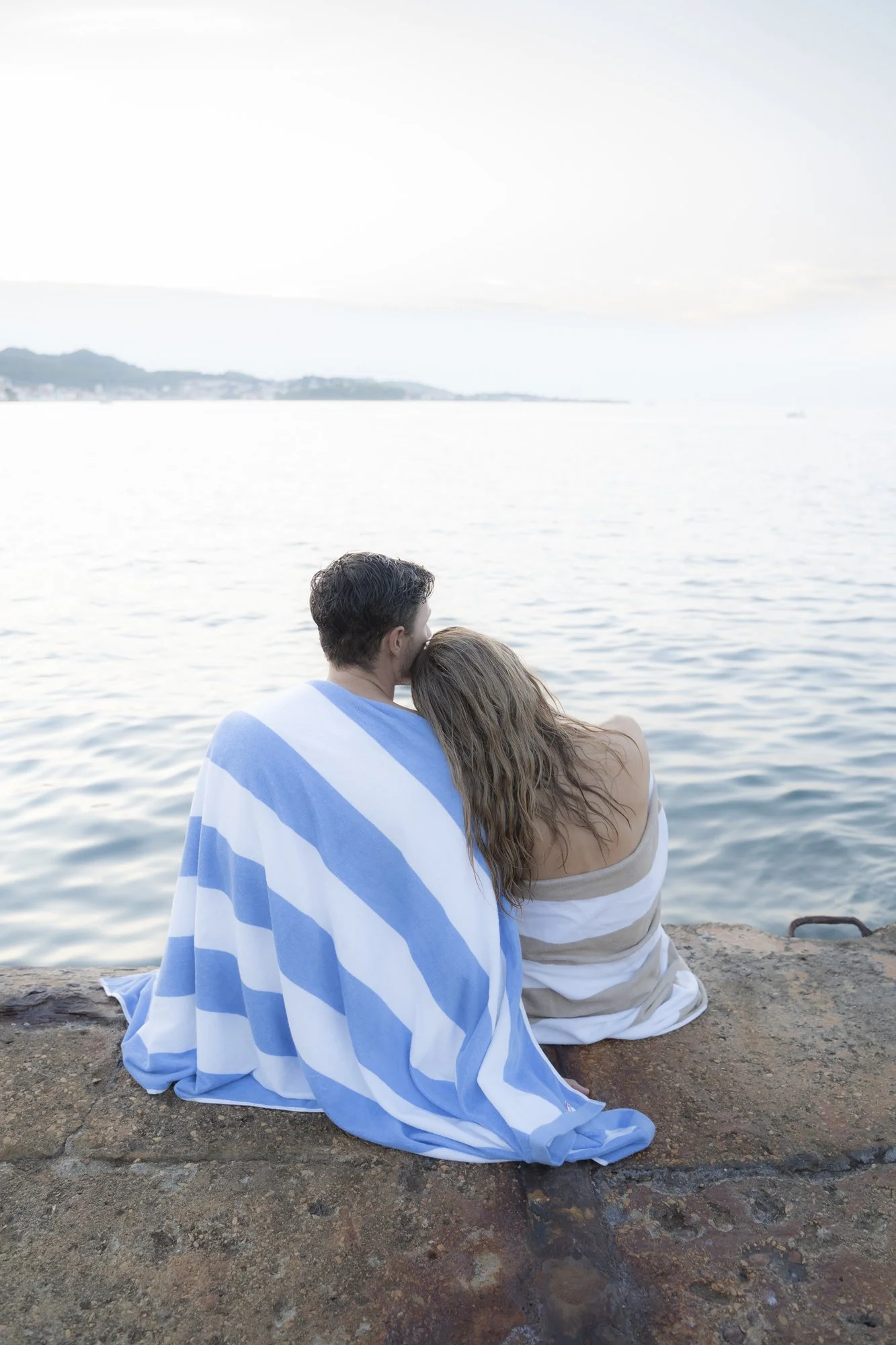A man and woman sitting by the water, wrapped in striped towels, enjoying a peaceful moment by the ocean or lake. Croatia, Zadar. Couple Photoshoot. Modern Wedding, modern couples