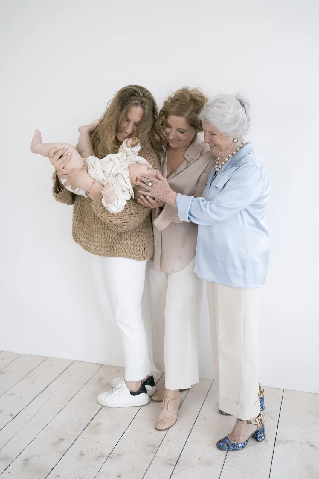 Four women, including a young woman, middle-aged woman, and an elderly woman, holding and playing with an infant. They are all smiling and standing against a plain white wall.