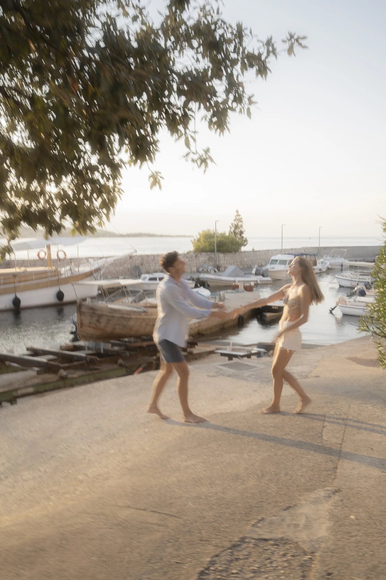 Two women dancing on a pier near boats and water, with trees and a clear sky in the background, during sunset.