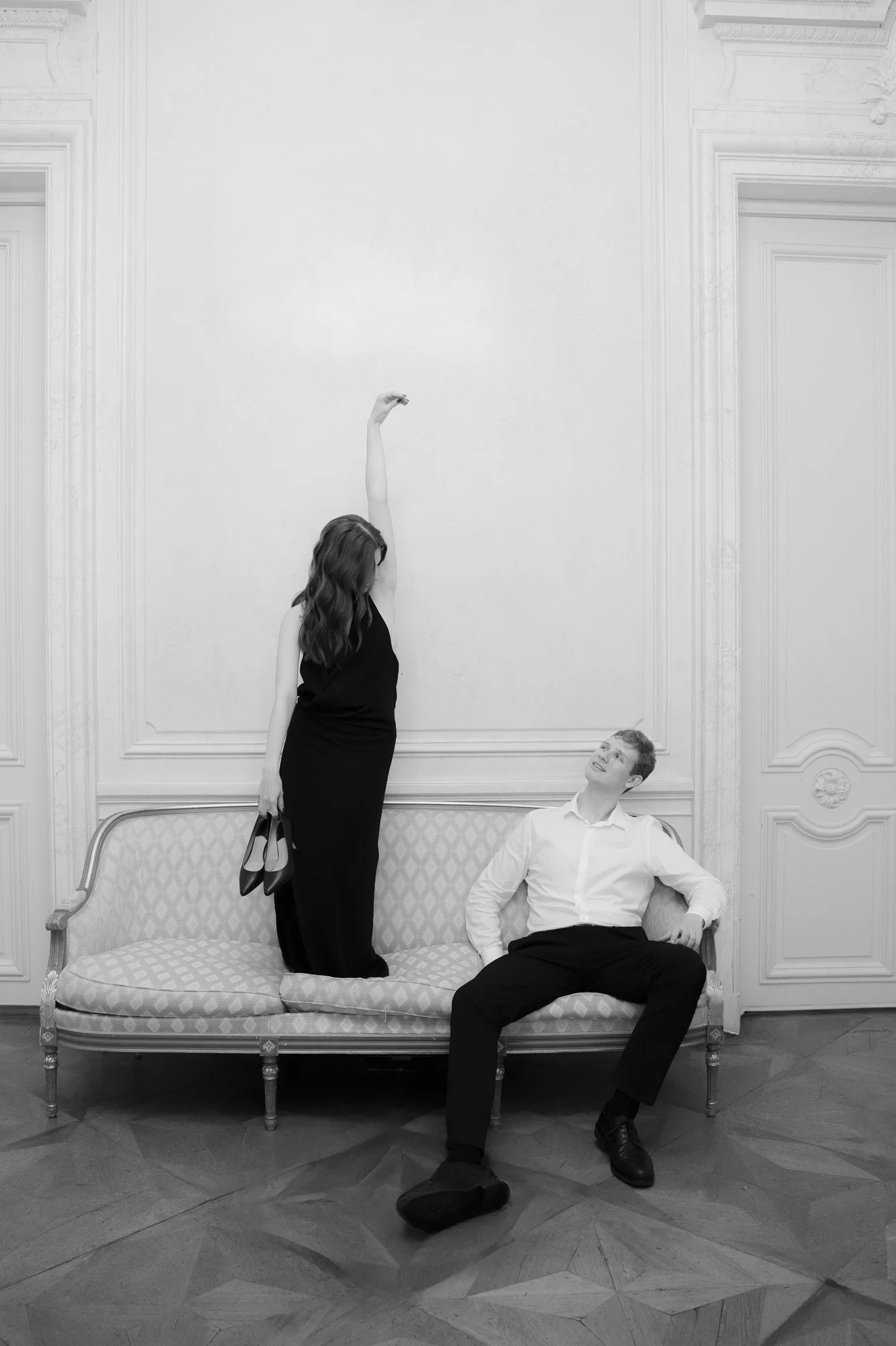 A black and white photo of a woman standing on a vintage couch, stretching her arm upward, and a man sitting on the couch looking up at her. The setting appears to be an elegant room with detailed wall paneling.