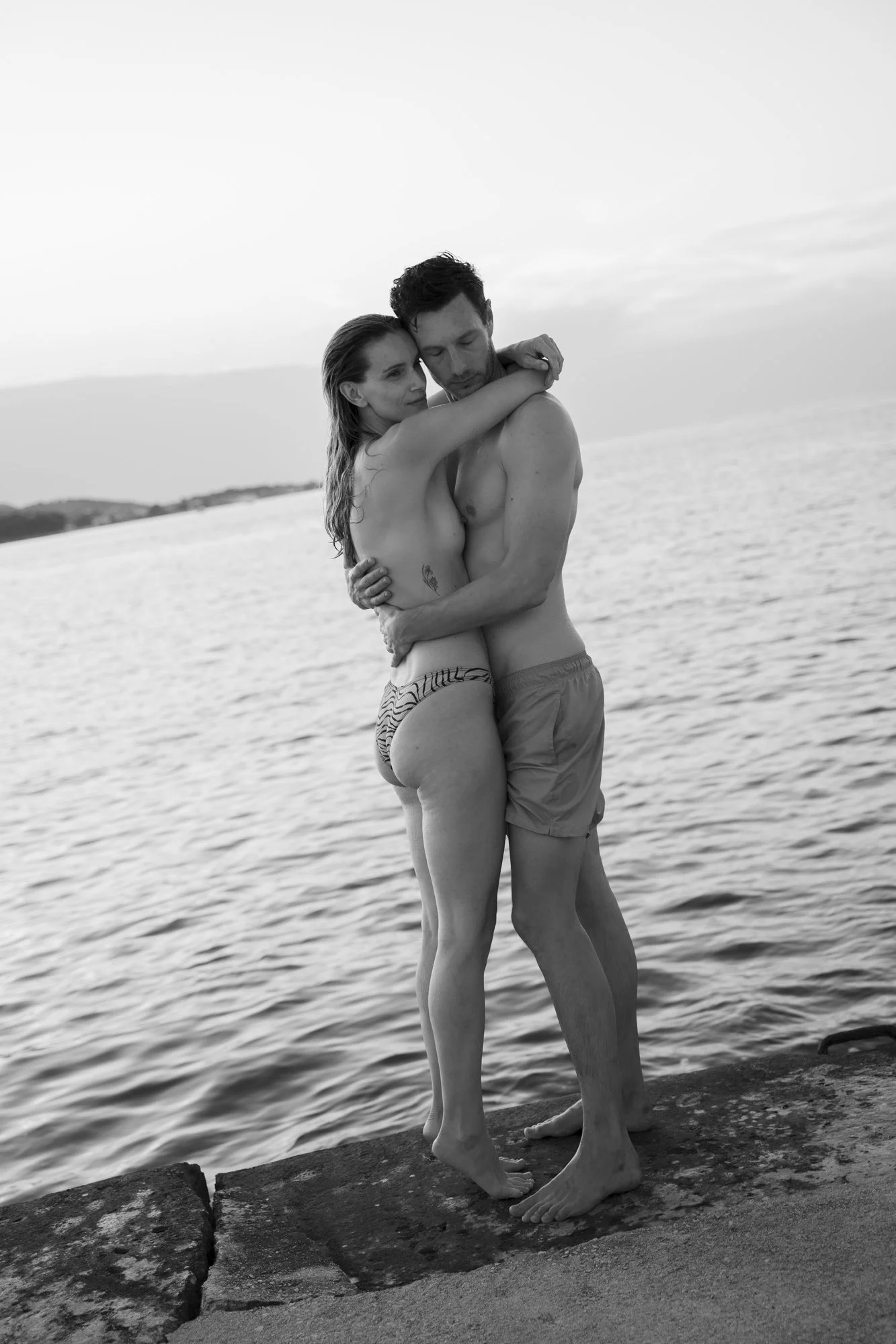 A black and white photo of a couple hugging by the water at the beach, embracing each other closely.