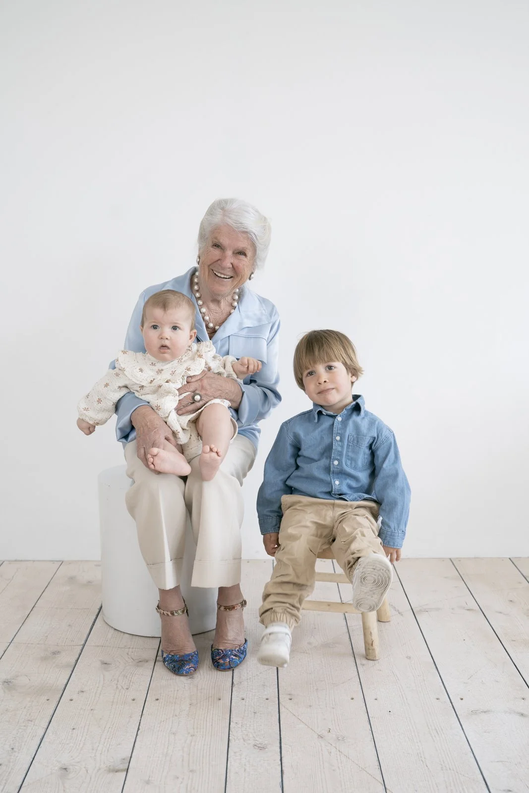 An elderly woman sitting on a white stool, smiling, holding a baby girl, with a young boy sitting on a small wooden chair beside her, all in front of a plain white background.