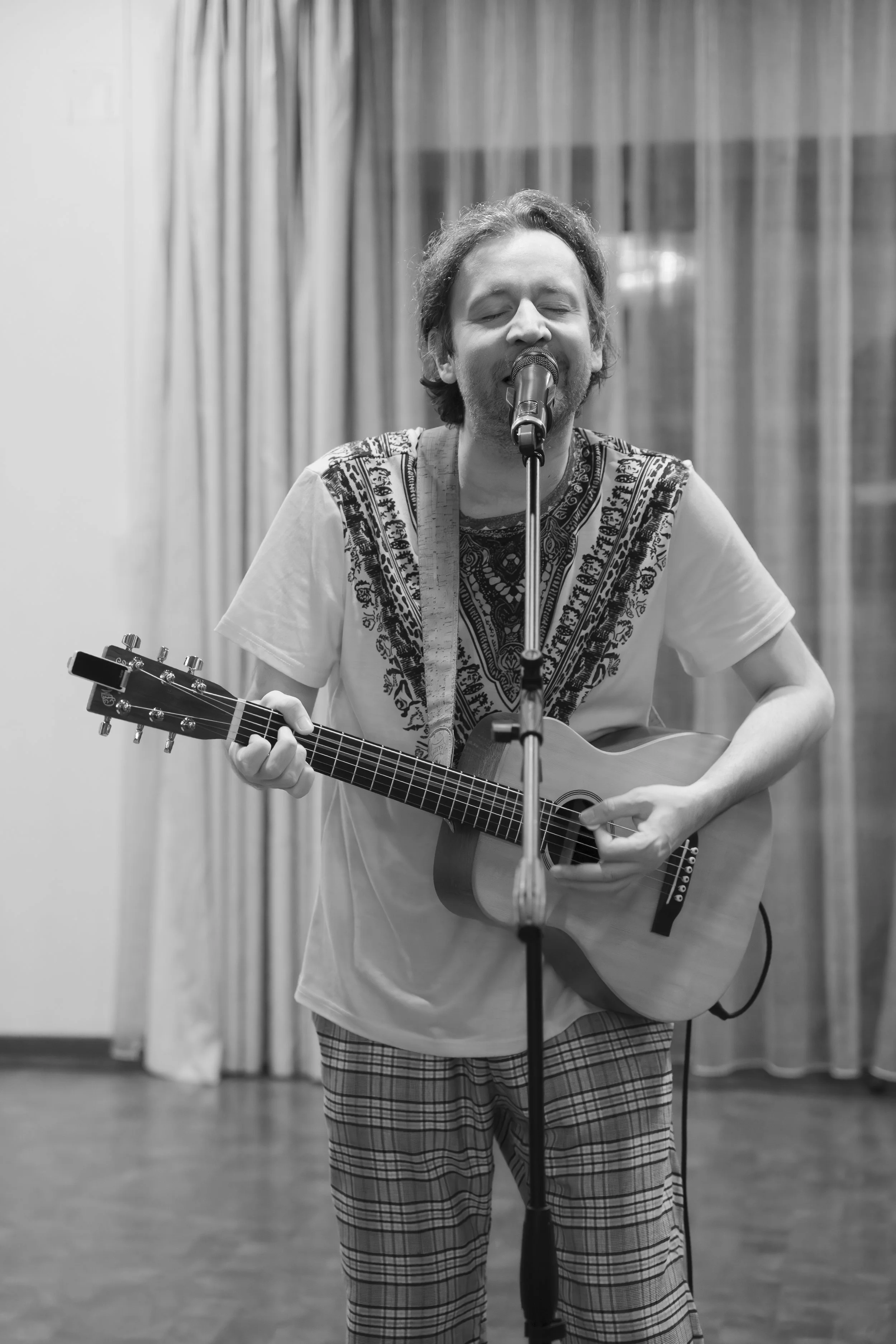 A man singing into a microphone while playing an acoustic guitar, with curtains in the background.