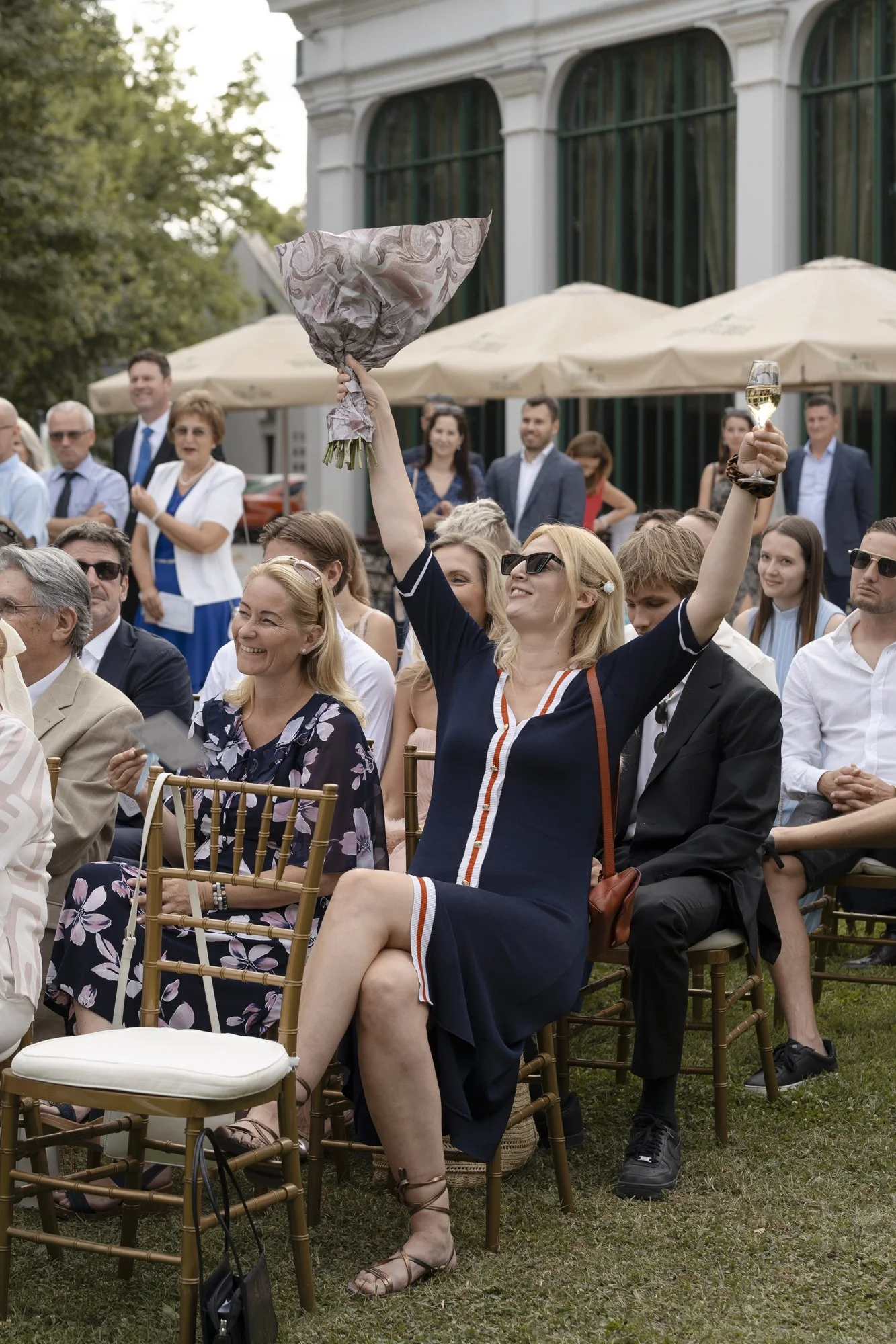A woman wearing sunglasses and a dark dress with white, orange, and navy stripes is sitting on a gold-colored chair at an outdoor event, smiling and raising a bouquet of flowers and a glass of champagne in celebration.