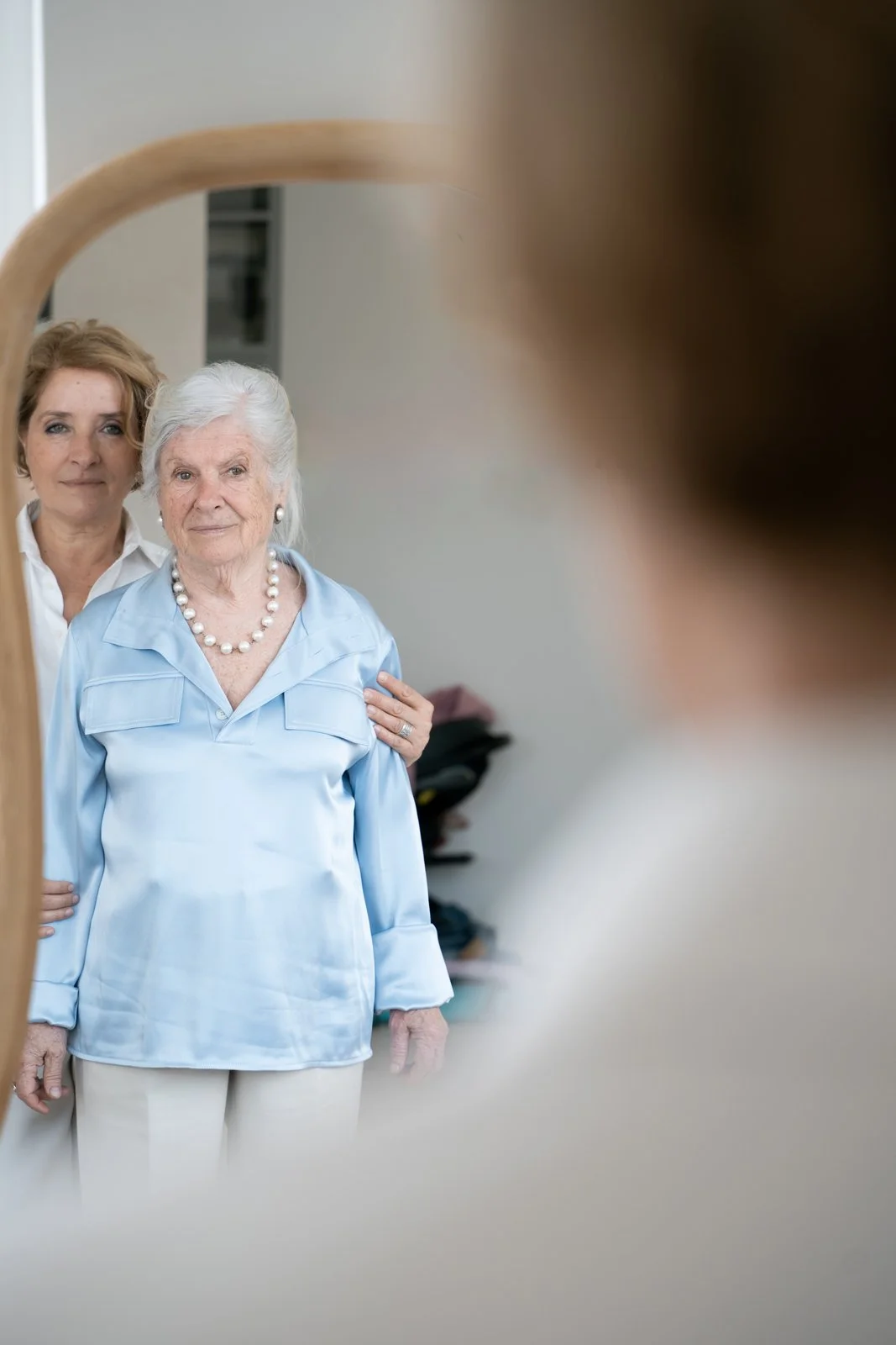 An elderly woman with white hair and a pearl necklace looks into a mirror, with a middle-aged woman behind her helping adjust her clothing. The mirror reflects their faces and upper bodies, and the scene appears to be in a home or dressing room.