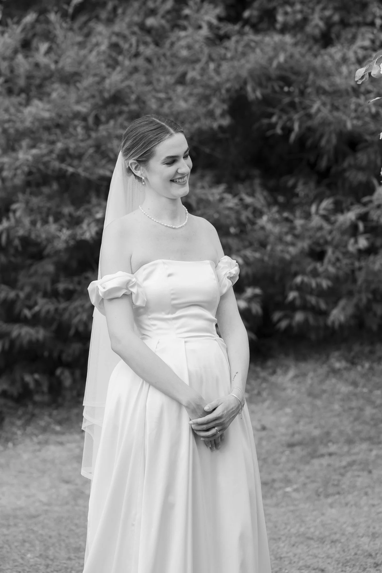 Black and white photo of a bride in an outdoor setting, wearing a strapless wedding gown and veil, smiling softly with hands clasped in front of her, surrounded by trees and foliage.