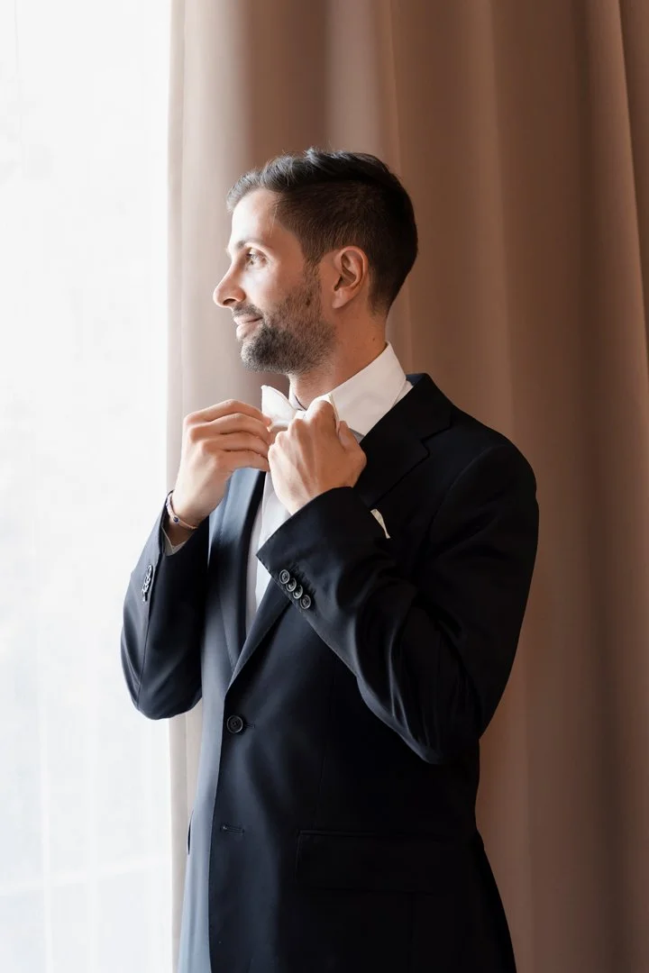 Man in a black tuxedo adjusting his white bow tie, standing near a window with curtains.