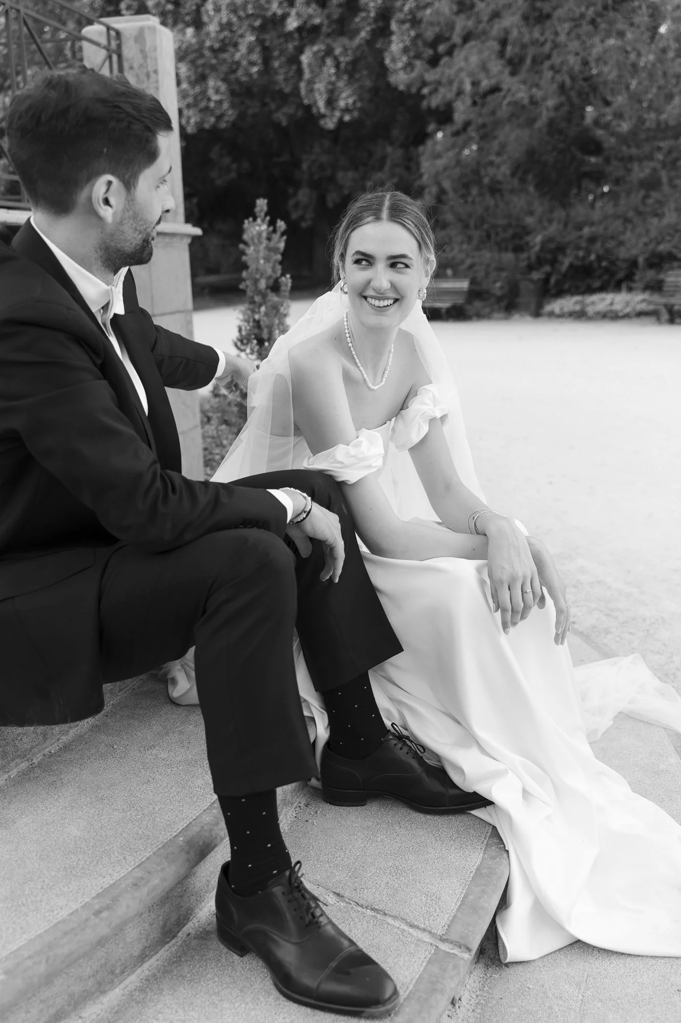 Black-and-white photo of a bride and groom sitting on steps outdoors, smiling at each other. The bride is wearing a wedding dress with puffed sleeves, a pearl necklace, earrings, and a veil. The groom is in a dark suit with a white shirt, and they are holding hands.