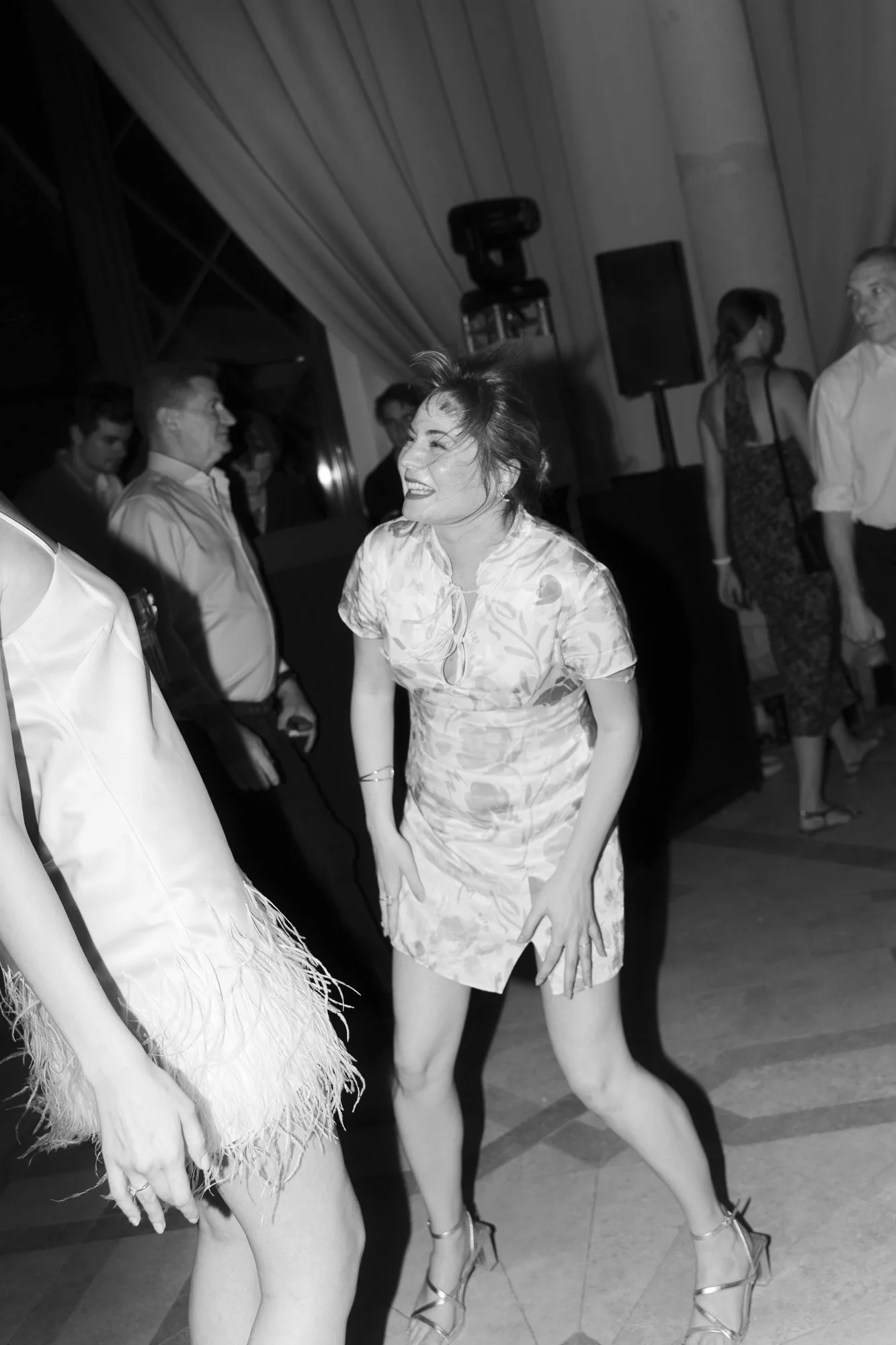 A woman dancing and laughing at a party or event. She wears a short dress with a floral pattern and high heels. Other people are in the background, some looking at her and others talking, in a room with curtains and stage equipment.