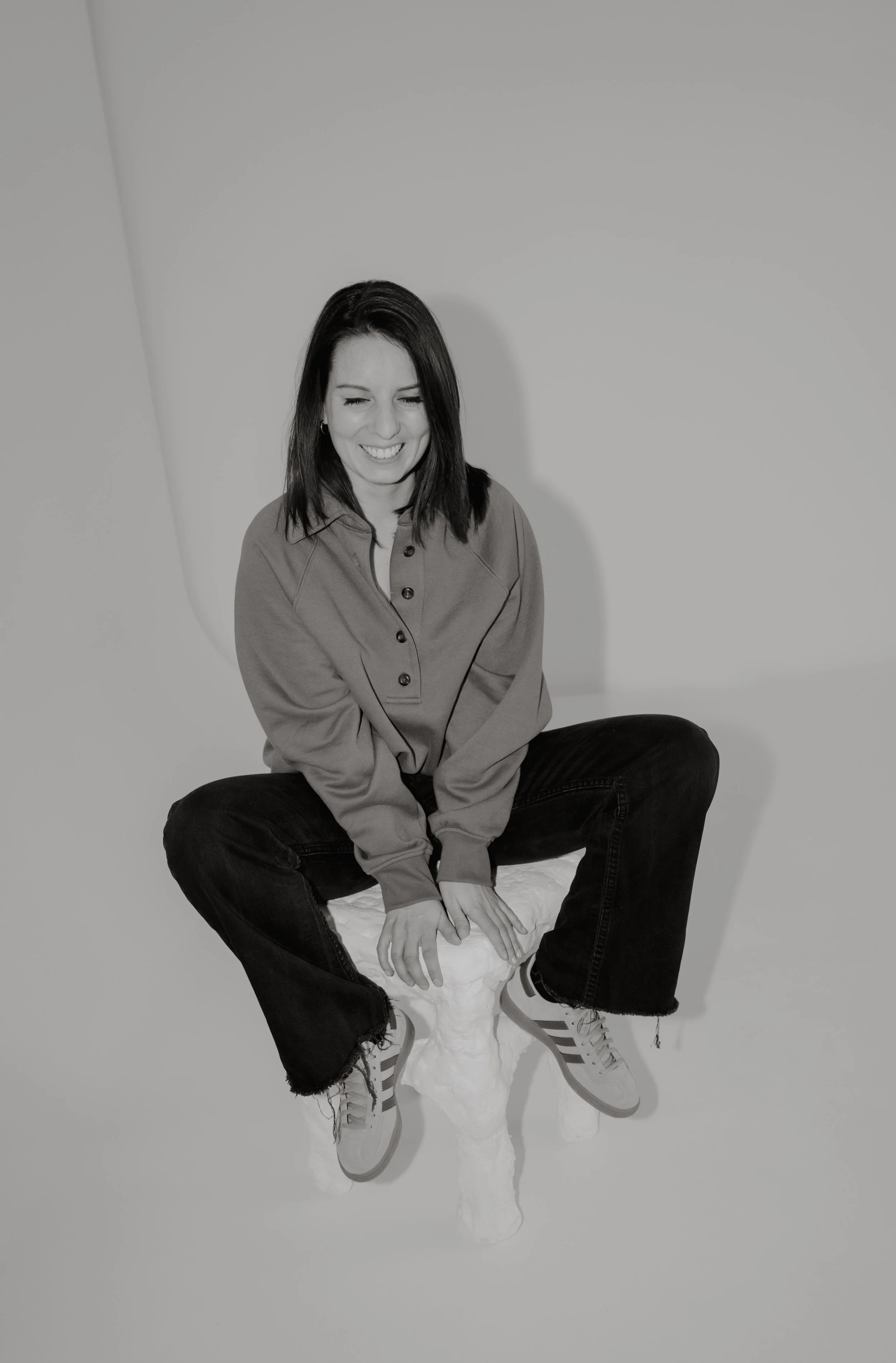 A woman sitting cross-legged on a white whimsical stool against a plain backdrop, smiling and laughing, wearing a loose shirt, jeans, and sneakers.