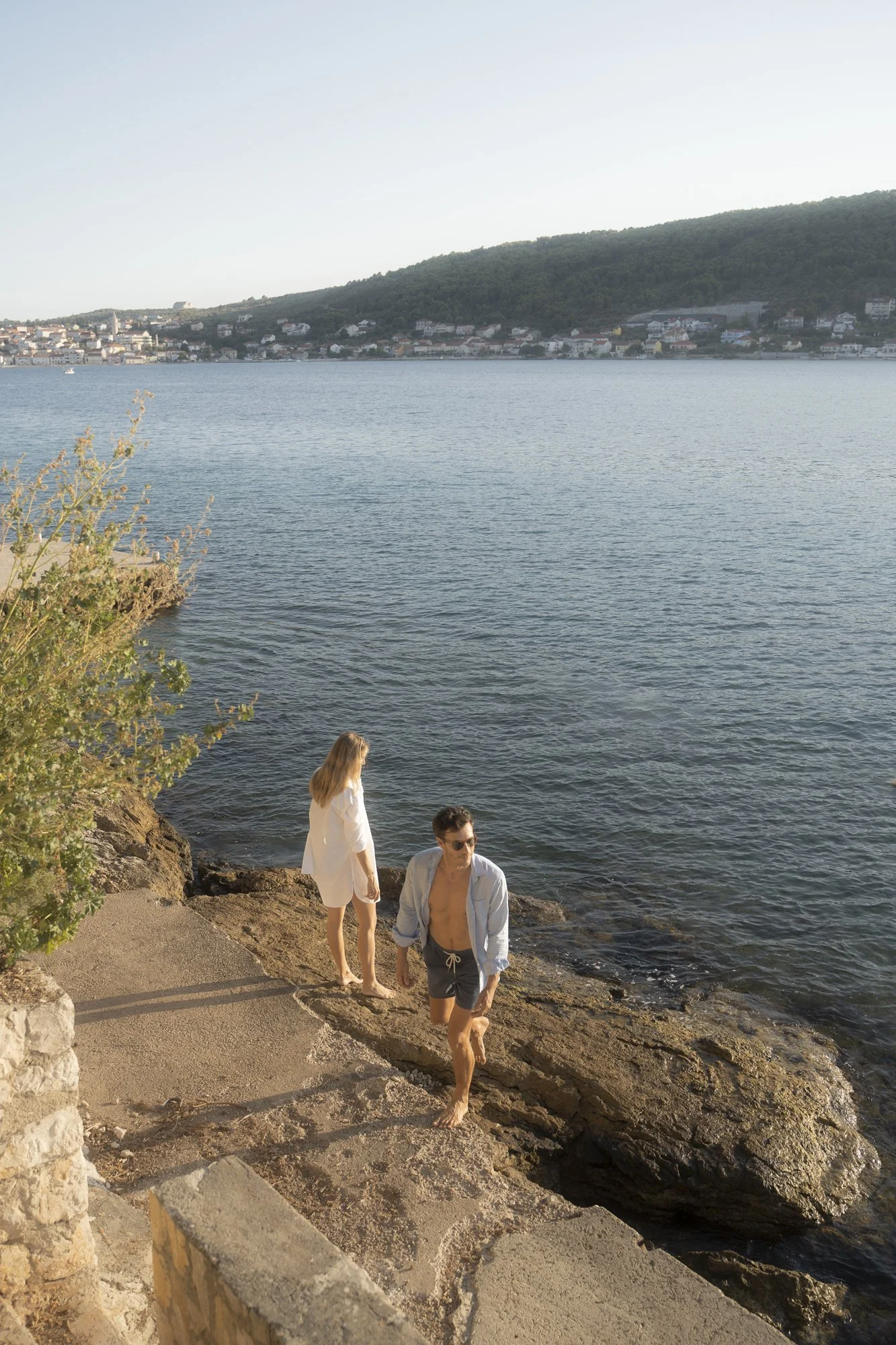 A man and a woman walk down rocky stairs to a body of water near a coastline with a hillside and houses in the background.