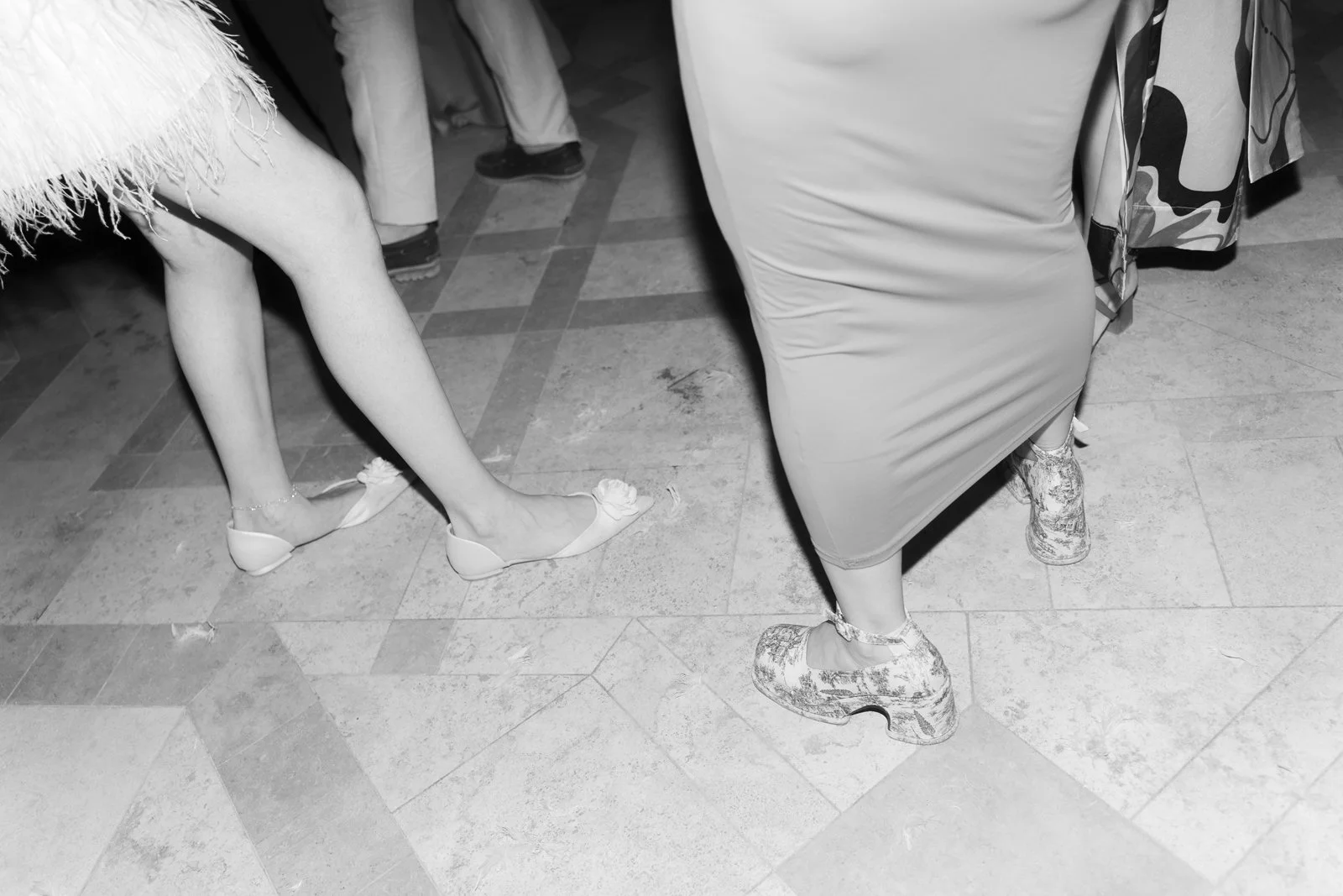 Close-up view of people's legs and shoes on a tiled floor, with some people sitting and standing around.