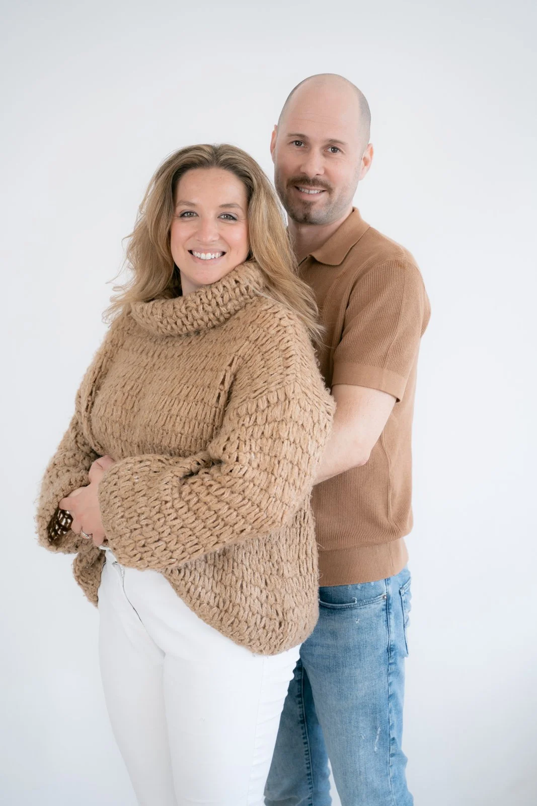 A smiling woman with blonde hair wearing a chunky beige sweater, standing in front of a smiling man with a shaved head, wearing a tan polo shirt. They are posing together against a plain white background.