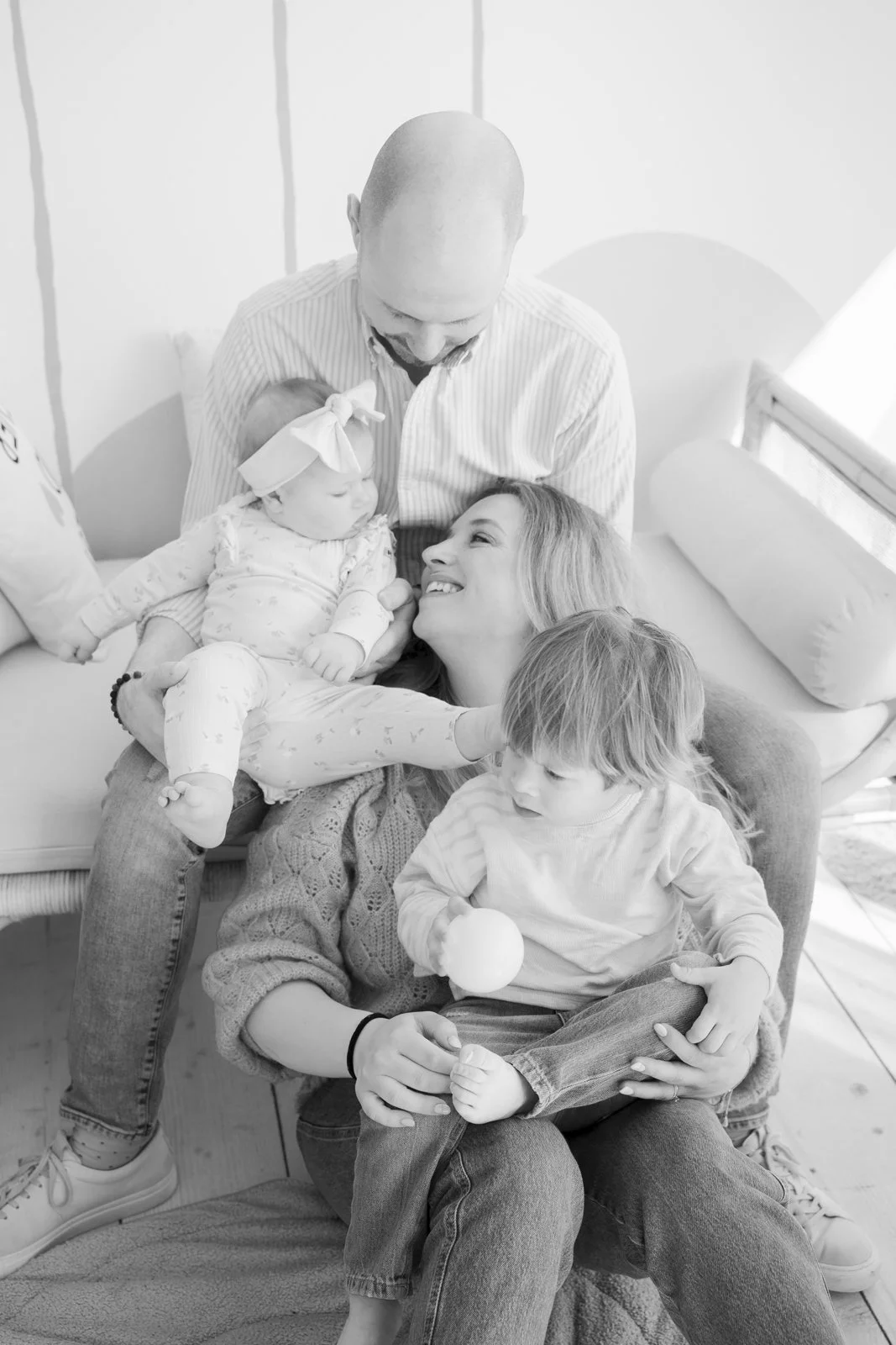 A family with two children, a woman, and a man in a hospital room. The woman is lying down, smiling at a baby girl on her lap. The man is looking down at the baby, and a young boy is sitting on the woman's lap holding an apple.