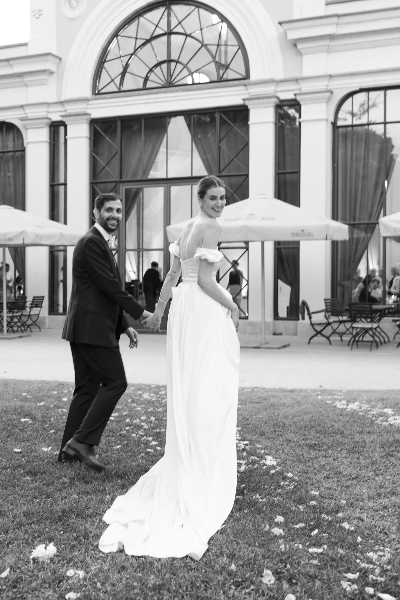 A bride and groom holding hands outdoors in front of a large building with large windows. The bride is smiling and looking back, wearing a wedding dress. The groom is also smiling, looking at the camera, and wearing a suit.