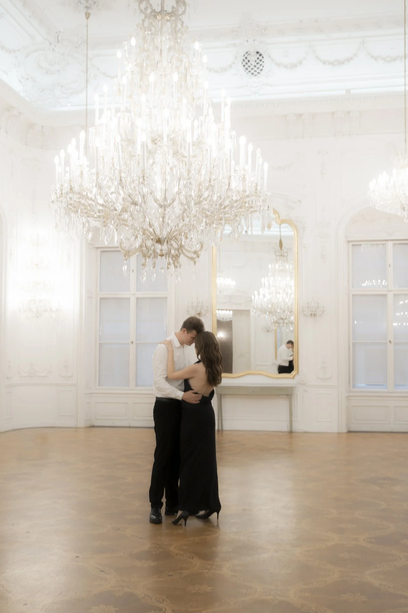 A couple dancing in a grand, elegant ballroom with ornate white walls, large windows, a gold-framed mirror, and a large crystal chandelier.