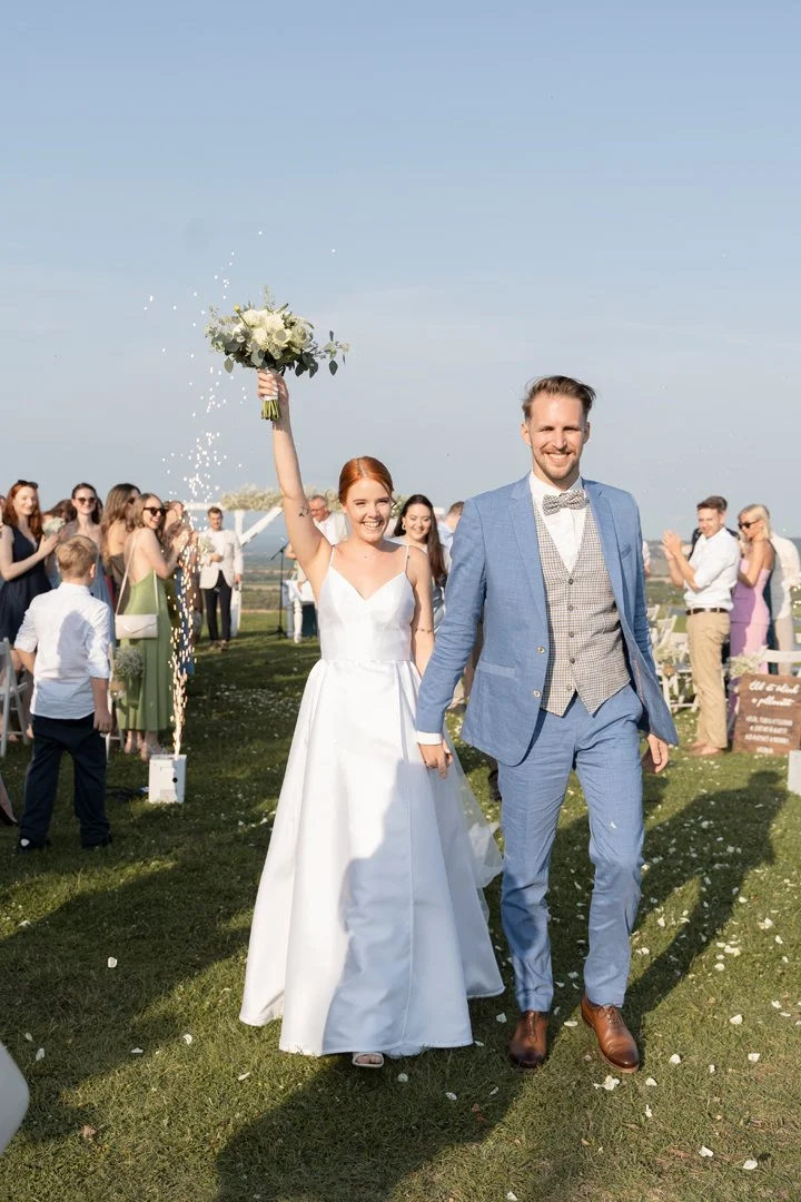 A newlywed couple walking hand in hand outdoors during their wedding celebration, with the bride holding a bouquet in the air and guests clapping and smiling in the background.