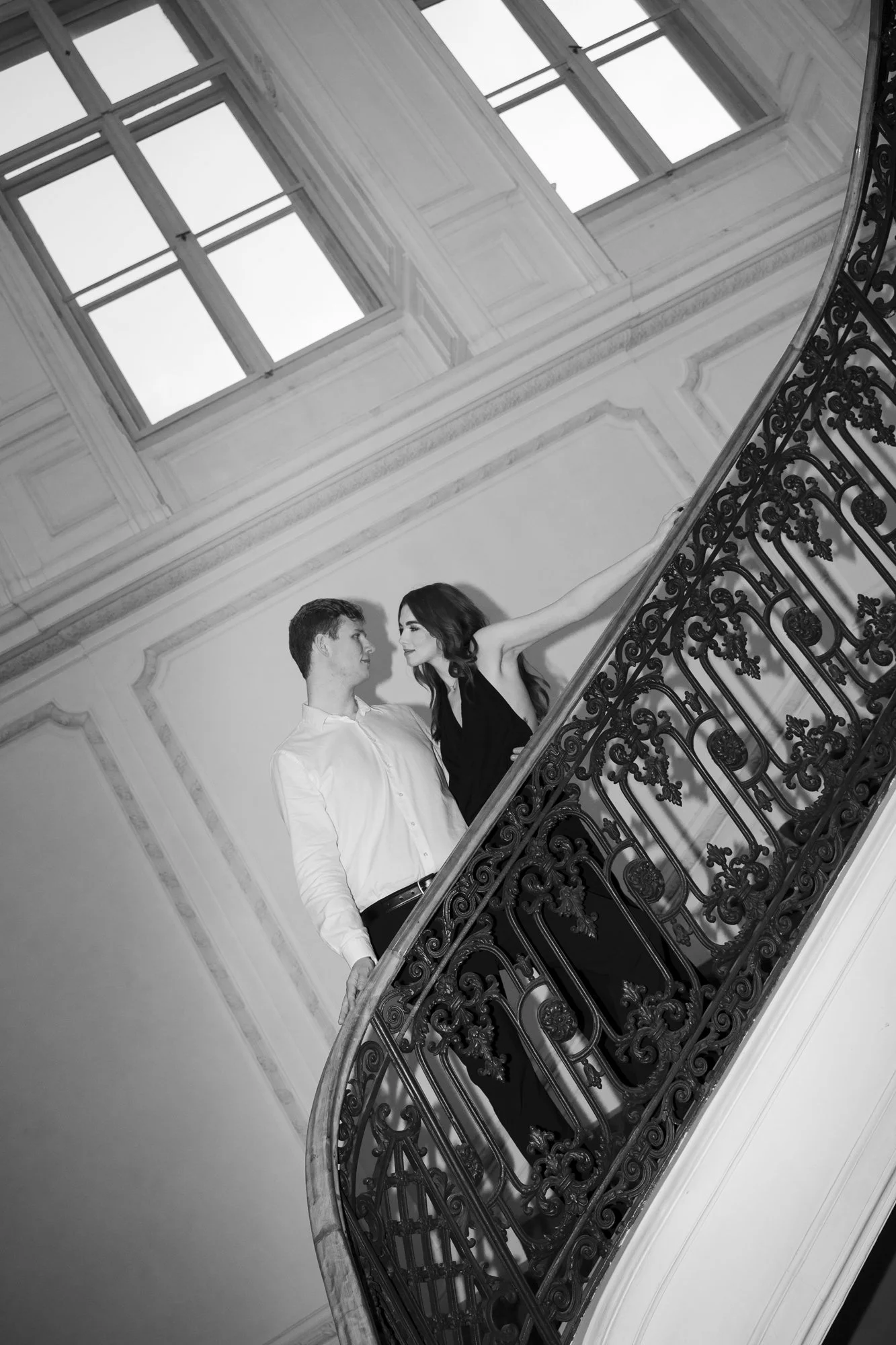 A black and white photo of a man and woman standing on a staircase with ornate wrought iron railing, gazing into each other's eyes, in a grand interior space with large windows and decorative molding.