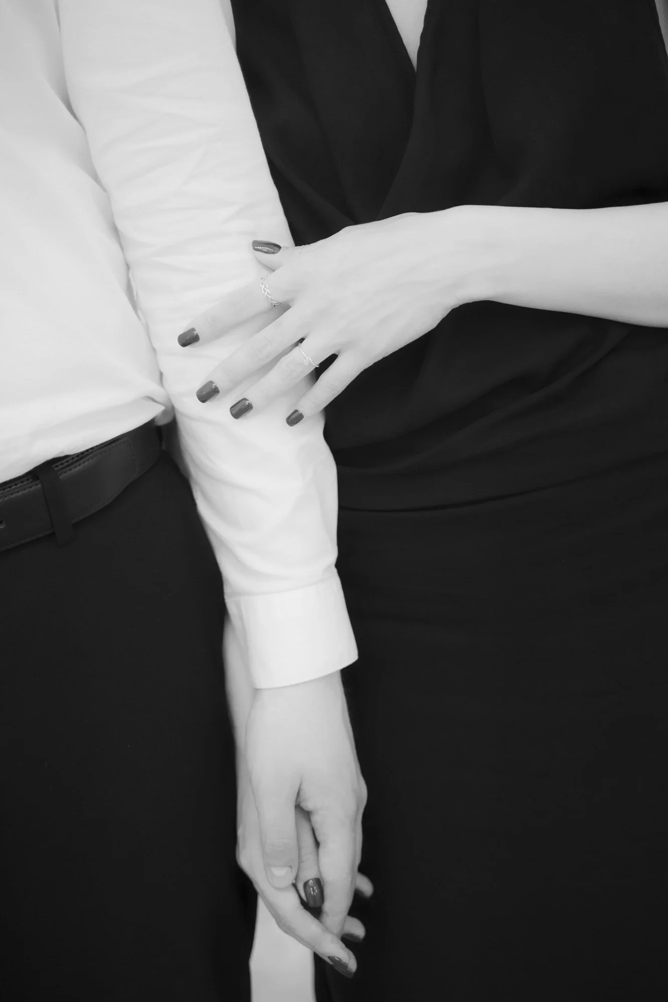 A close-up of a couple holding hands, with the woman's hand resting on the man's arm. The woman is wearing rings and has dark painted nails, while the man is dressed in a white shirt and black pants.