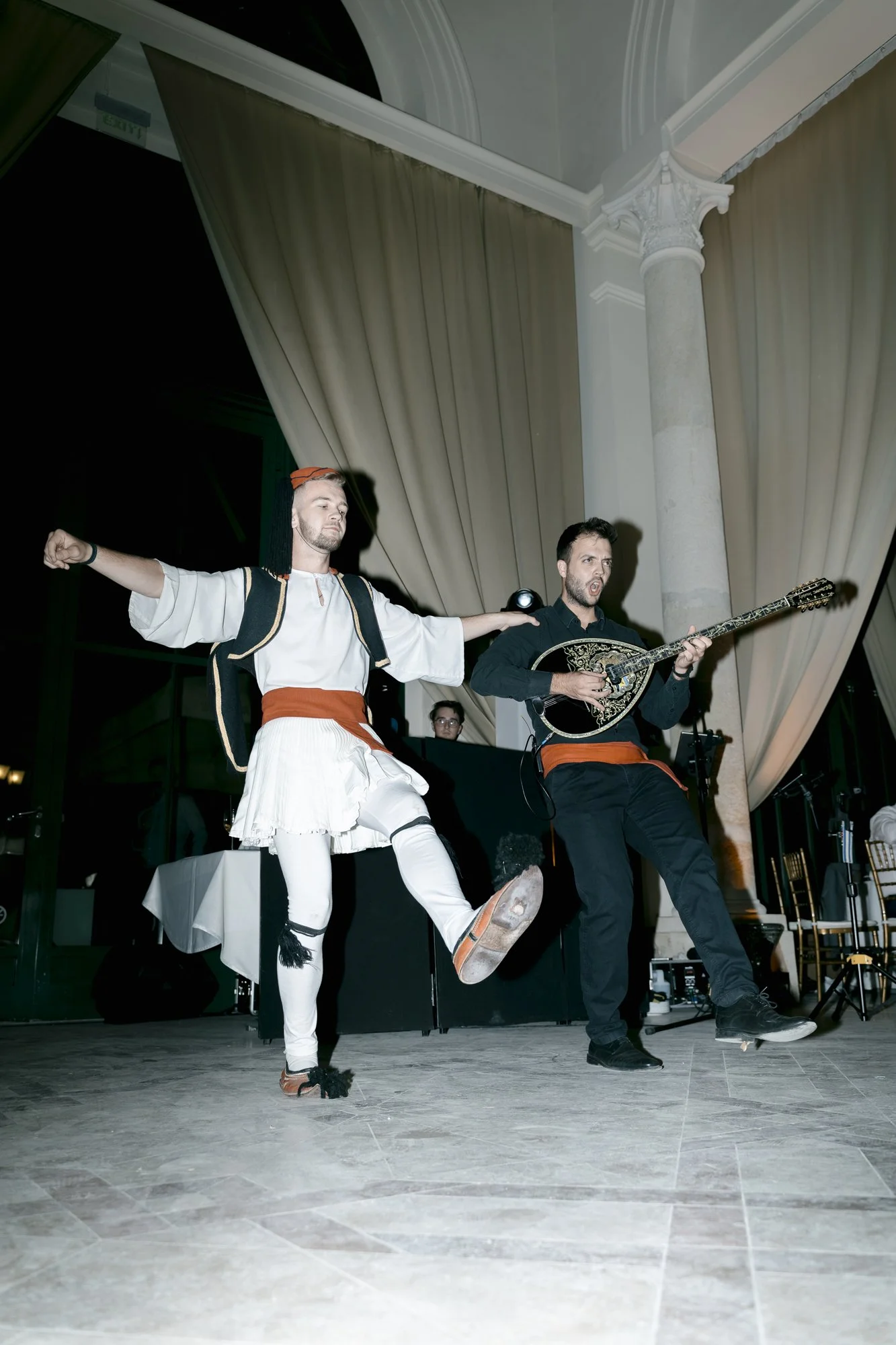 Two male performers in traditional Greek attire dancing and singing with a musician playing a stringed instrument on stage in an elegant hall with grand curtains and columns.