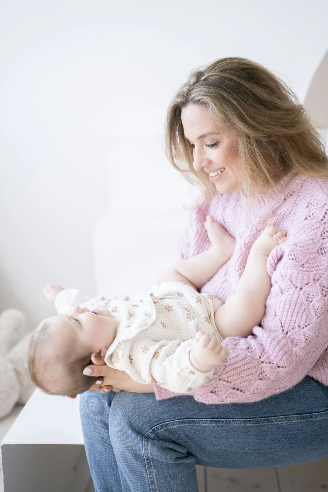 A woman in a pink sweater sitting on a white bench and holding a smiling baby girl in her arms, indoors with a white background.