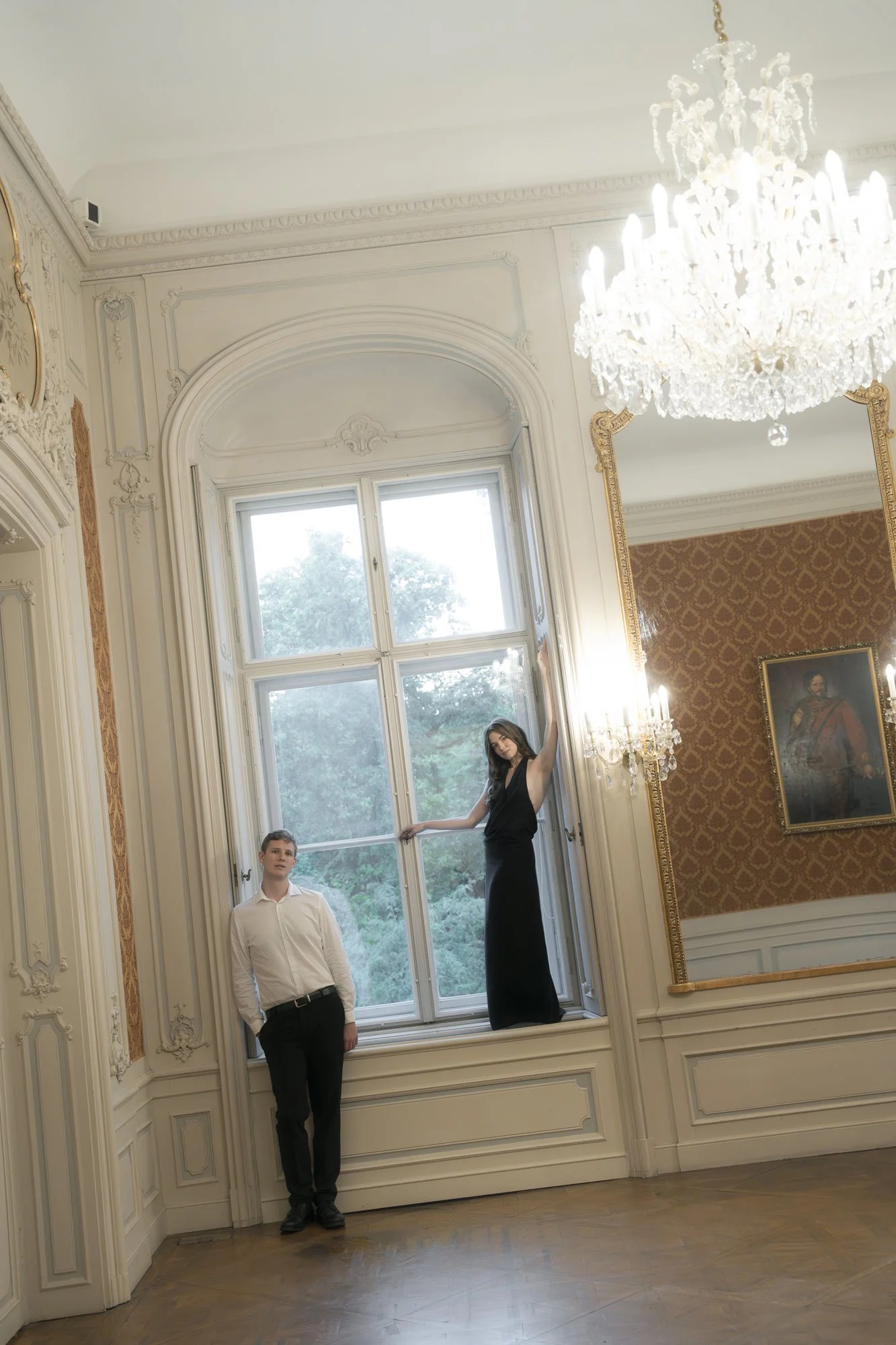 A young man in a white shirt and black pants stands next to a large window with a woman in a black dress sitting on the windowsill, in an elegant room with ornate white walls, golden accents, a chandelier, and a portrait painting.