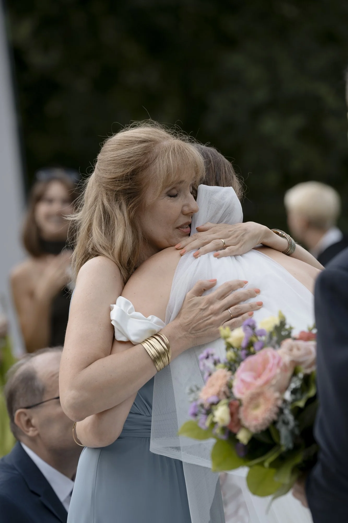 A woman with red hair wearing a light blue dress embraces a person in a white dress with a veil. The woman has her eyes closed and is holding a bouquet of flowers, and there are people in the background at an outdoor event.
