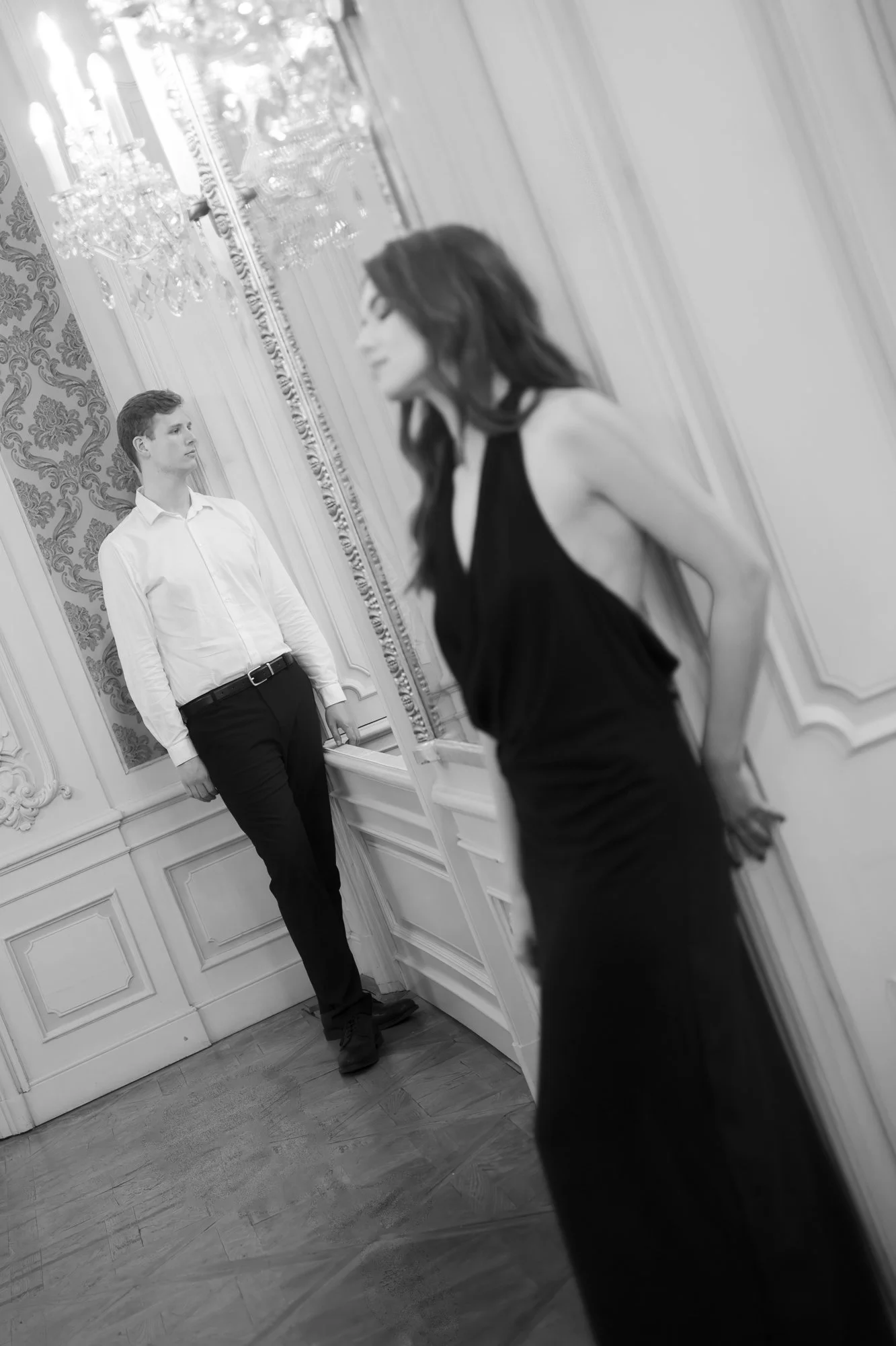 A black-and-white photo shows a woman in a black dress standing with her back against a wall, her hands behind her back, gazing at a man in a white shirt and black pants who is standing and looking at her in an ornately decorated room.