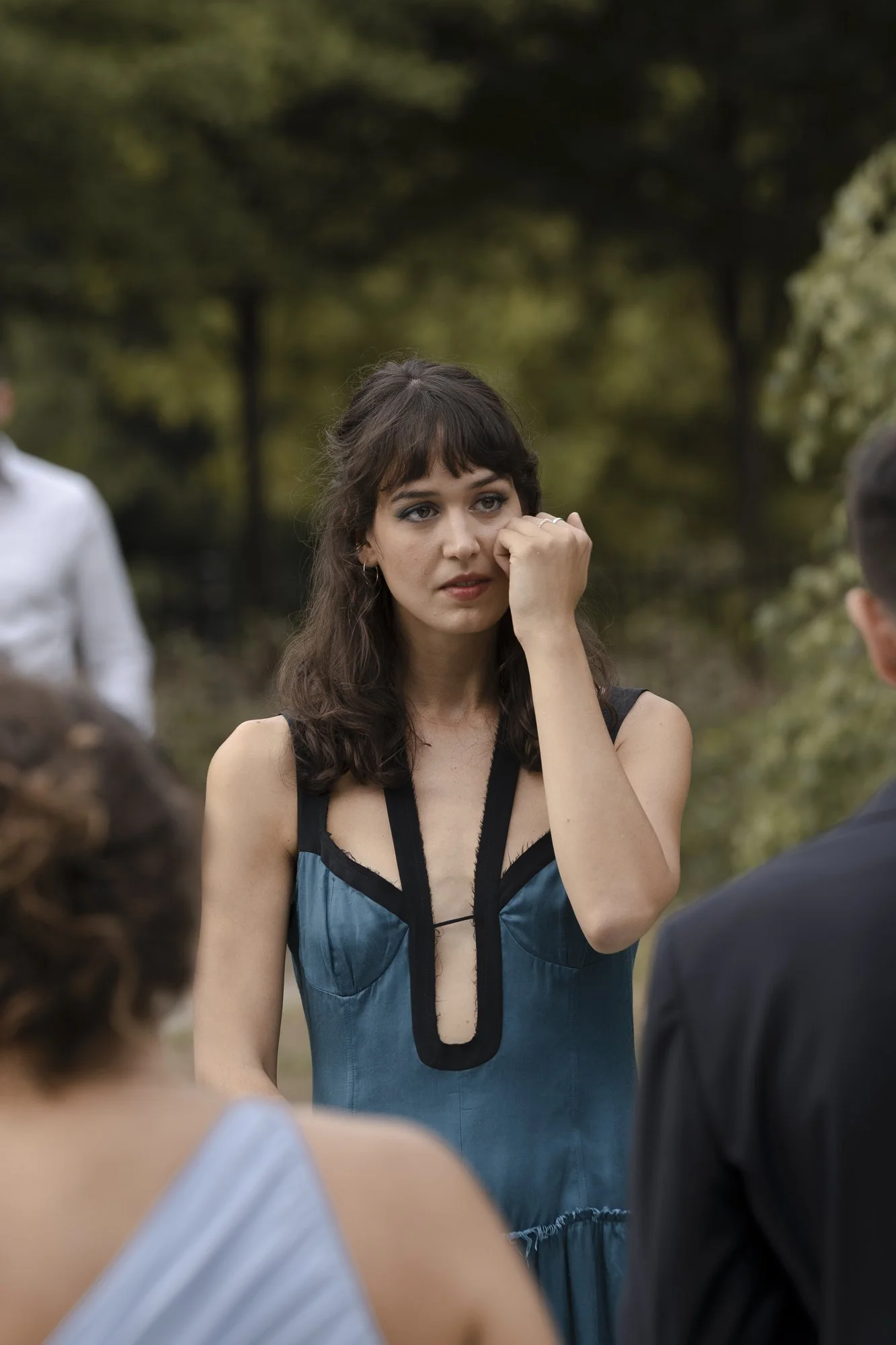 A young woman with brown hair and bangs wearing a blue dress with black accents is wiping her eye during an outdoor event, possibly a wedding, with several people around her in a natural setting with trees.