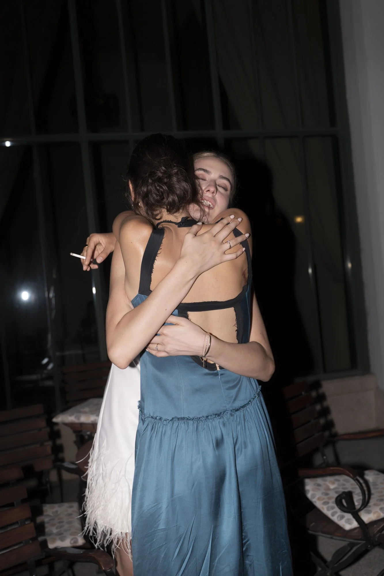 Two women hugging each other tightly, one crying with eyes closed at night indoors, one holding a cigarette.