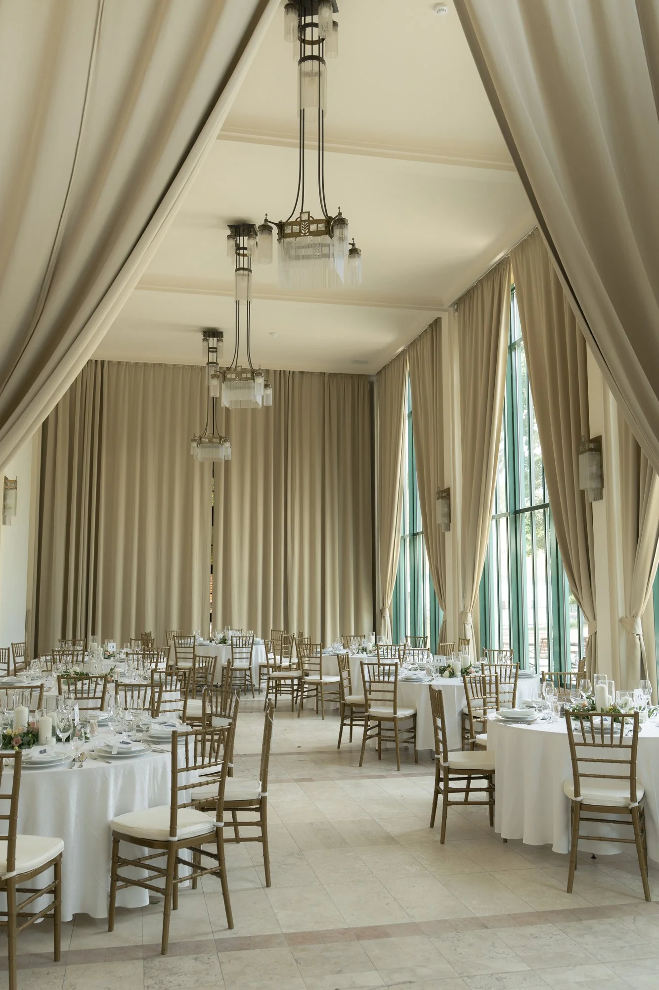An elegantly decorated event hall with round tables covered in white tablecloths and surrounded by wooden chairs. The hall features large windows with green frames, tall cream-colored curtains, and modern hanging light fixtures. The floor is tiled, and the tables are set with glassware, plates, and floral centerpieces.