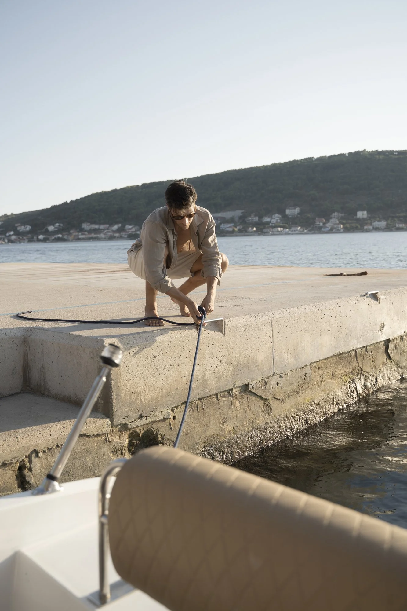 Man crouching on pier, adjusting boat line with water and hills in background.
