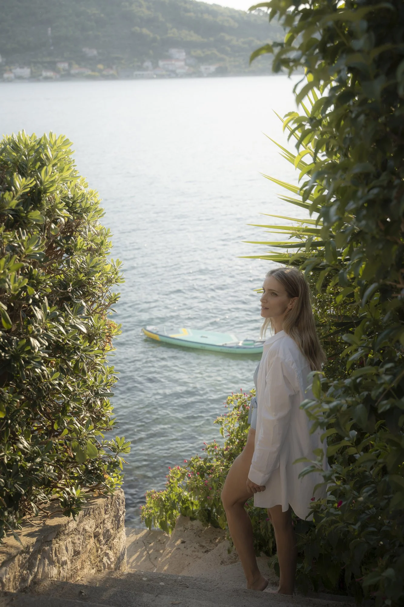 A woman in a white shirt stands on a stone step among lush green bushes, overlooking a body of water with a paddleboard floating nearby.