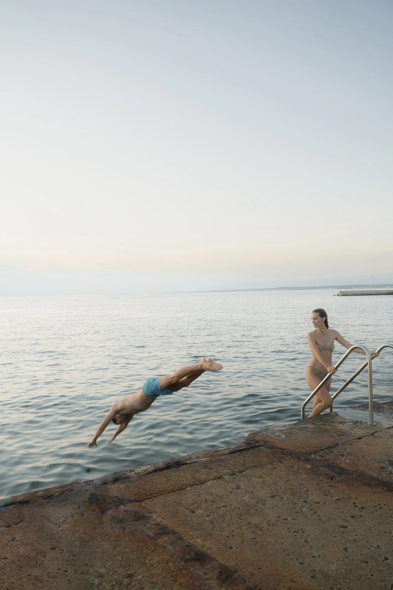A woman in a bikini is climbing out of the water on a metal ladder at a pier, while a man in swim trunks is diving into the water from the pier during sunset.