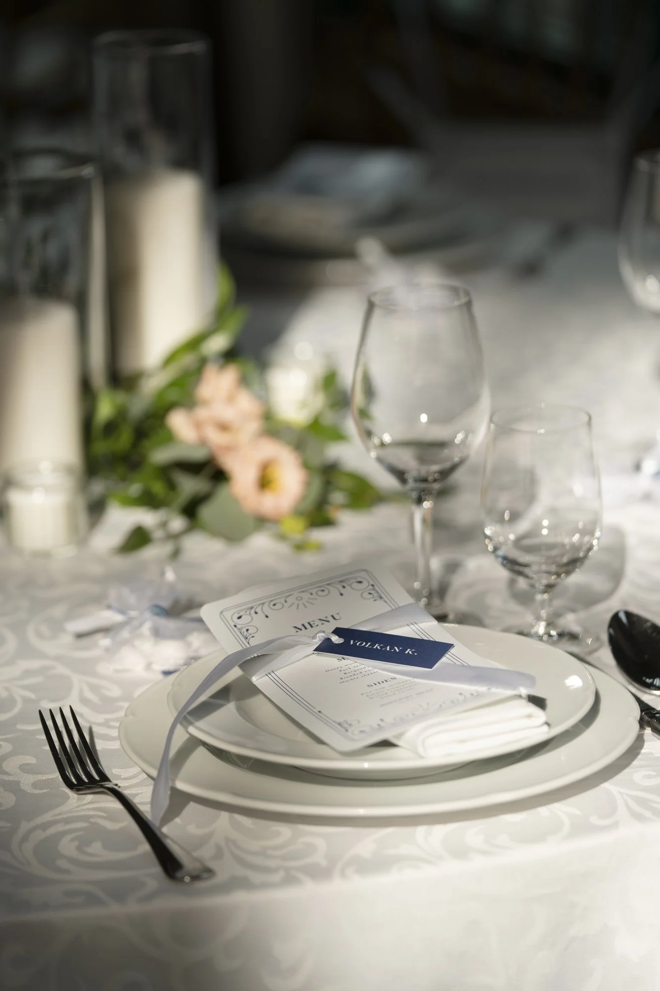Elegant dining table setting with a folded menu, a name card labeled 'Volkan K.', white plates, silverware, glasses, and a flower arrangement with candles.