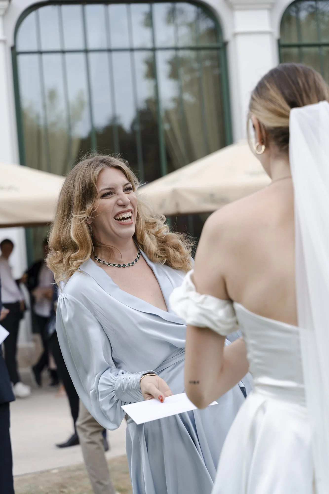 A woman with blonde hair and a blue dress smiling and handing a piece of paper to a woman in a white wedding dress outdoors.