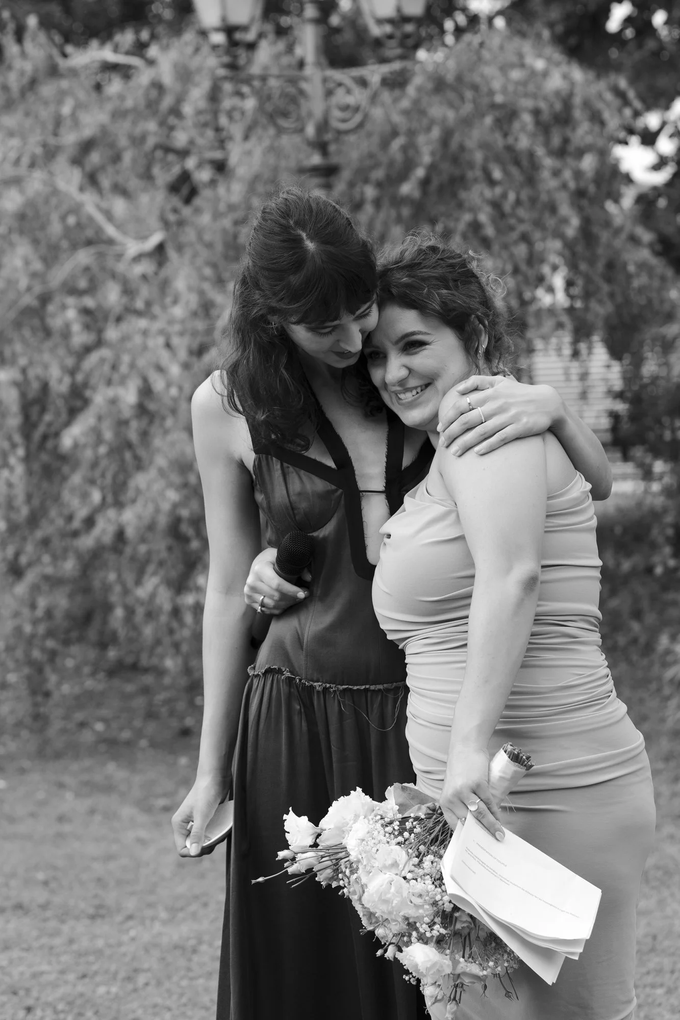 Two women hugging outdoors, one holding a bouquet of flowers and a rolled-up piece of paper, smiling.