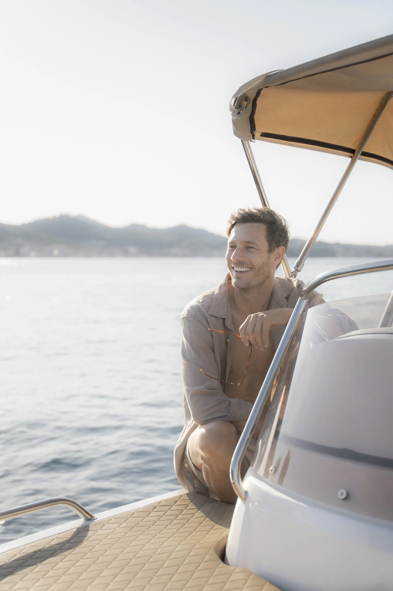 A man sitting on the deck of a boat, holding sunglasses, with a smile, near a body of water with distant hills in the background.