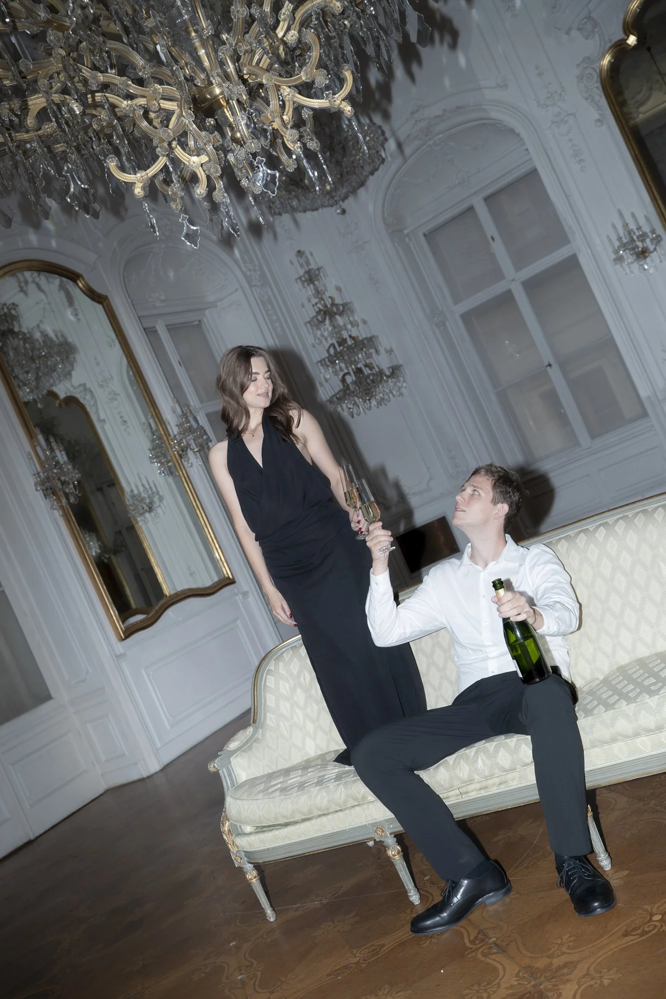A man sitting on a vintage cream-colored sofa with gold accents, holding a bottle of champagne in one hand and a glass in the other, looking up at a woman standing next to him. The woman is wearing a black dress, holding a champagne glass, and smiling. The room has ornate white walls with decorative moldings, large windows, and fancy chandeliers hanging from the ceiling.