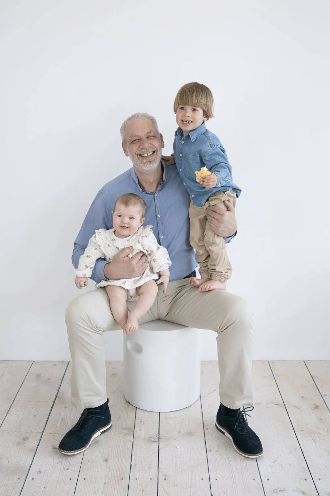 An elderly man sitting on a white cylindrical stool, smiling with two children, a toddler girl on his lap and a young boy standing on the stool. Modern family photoshoot.