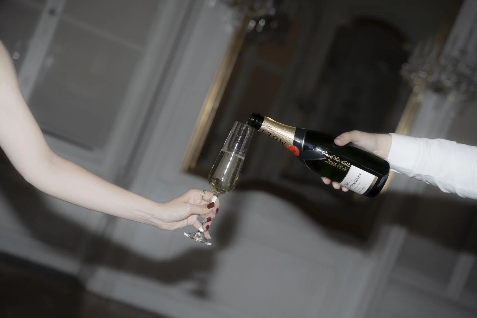 Someone is pouring champagne from a bottle into a champagne flute held by a woman with manicured nails.