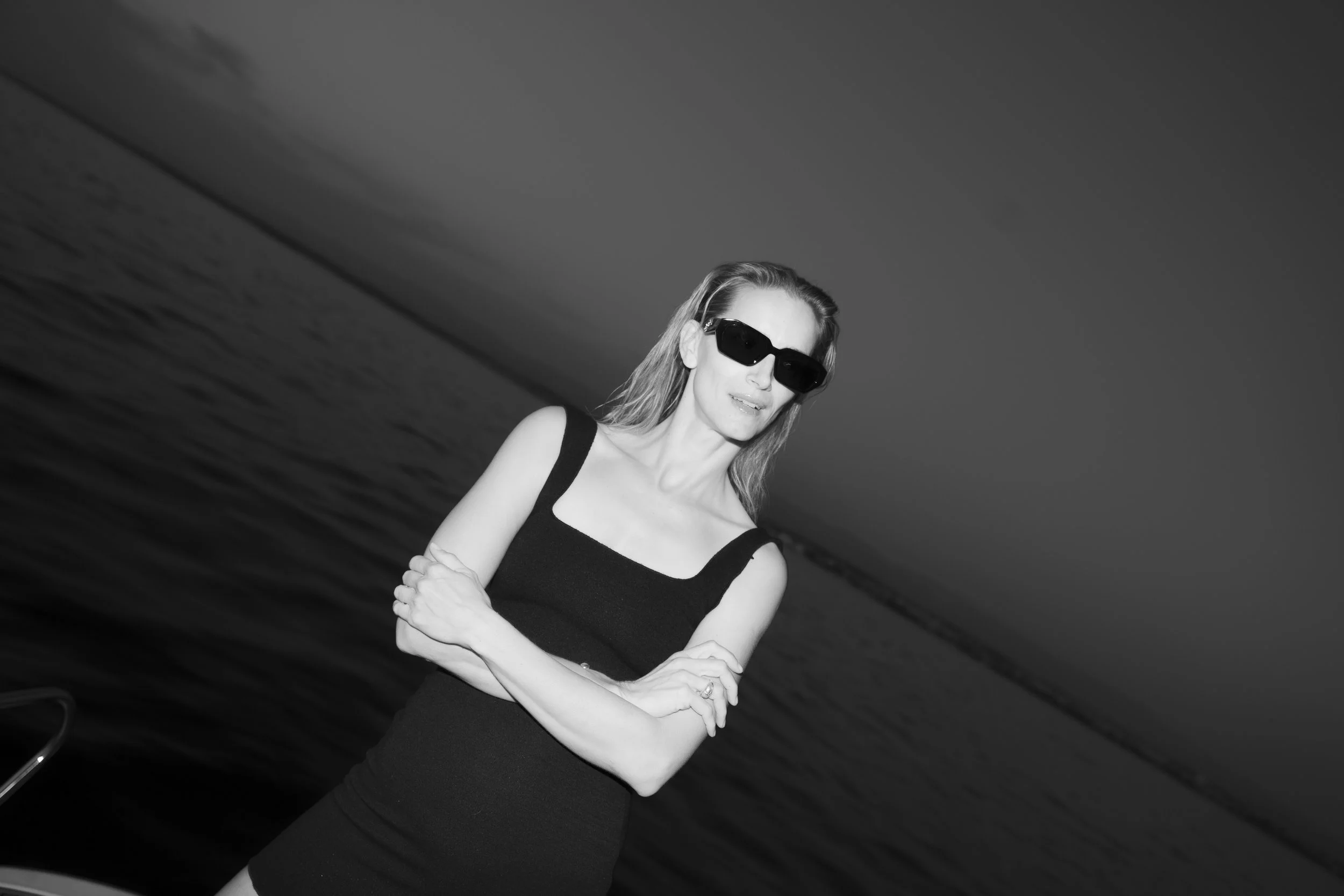 Black and white photo of a woman wearing sunglasses and a sleeveless black dress, standing near the water with her arms crossed.