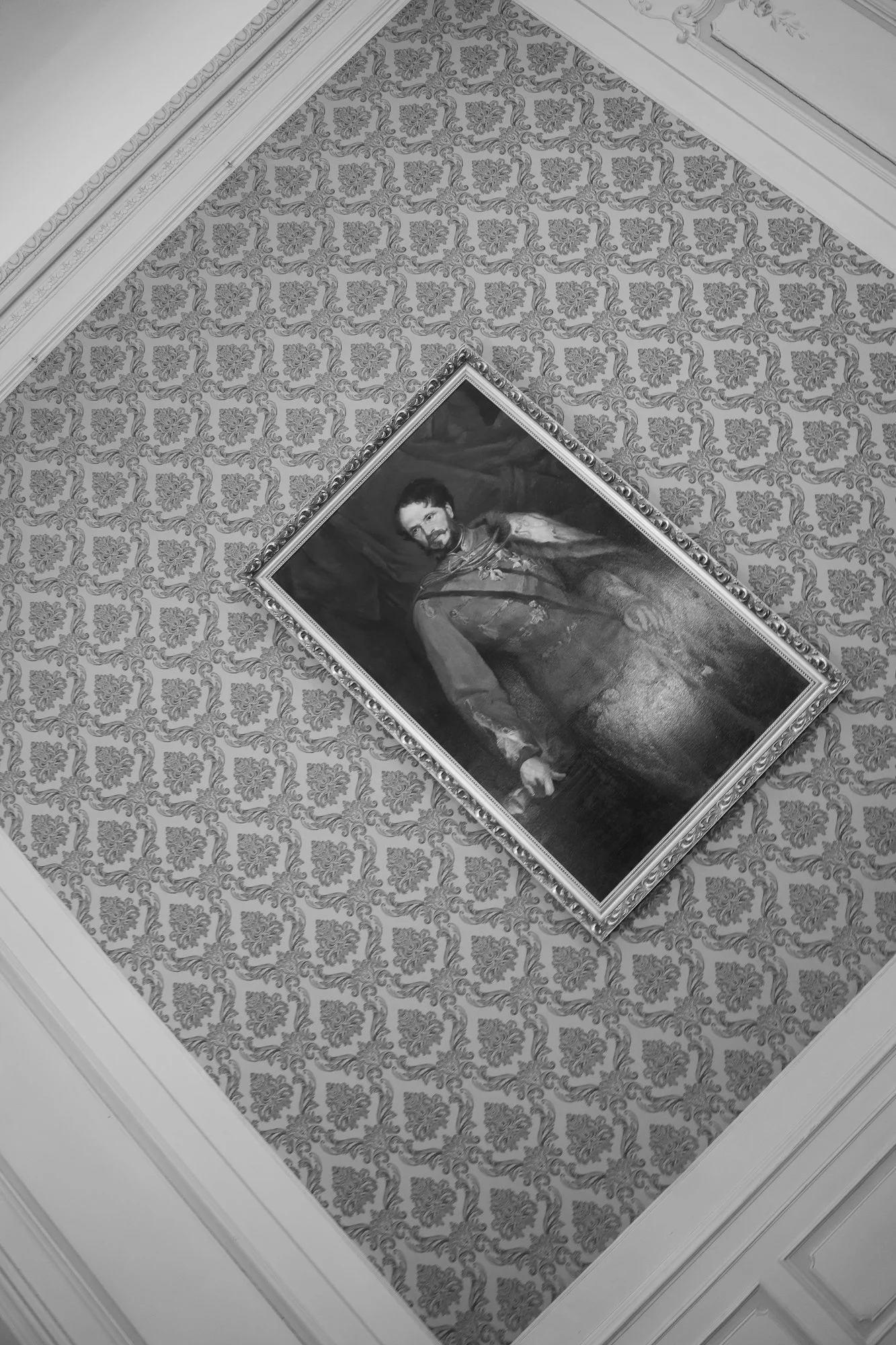 Black and white photograph of a man in a historical military uniform, hanging on an ornate, patterned wallpapered wall.