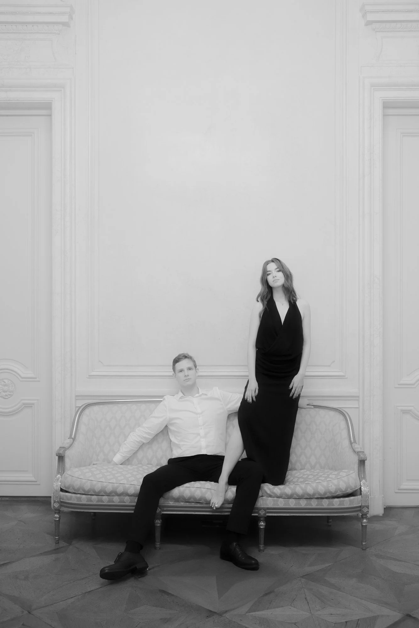 A man seated on a vintage sofa with an arm resting on an elaborate room wall, and a woman standing with her foot on the man's lap, both wearing formal black and white outfits, in an elegant room with ornate wall moldings.