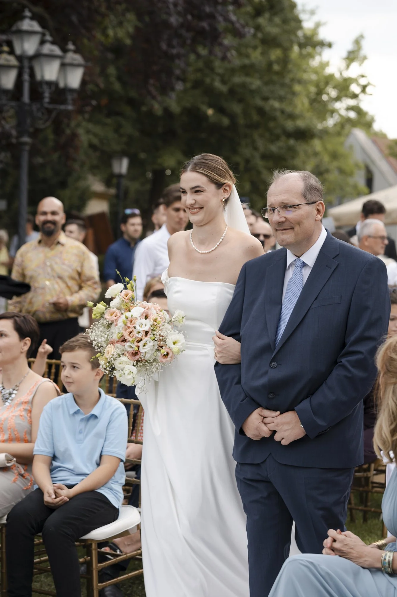 Bride in a strapless white wedding gown holding a bouquet of pink and white flowers, walking arm-in-arm with a man in a navy suit at an outdoor wedding ceremony, surrounded by seated guests and standing onlookers, with greenery and outdoor lights in the background.