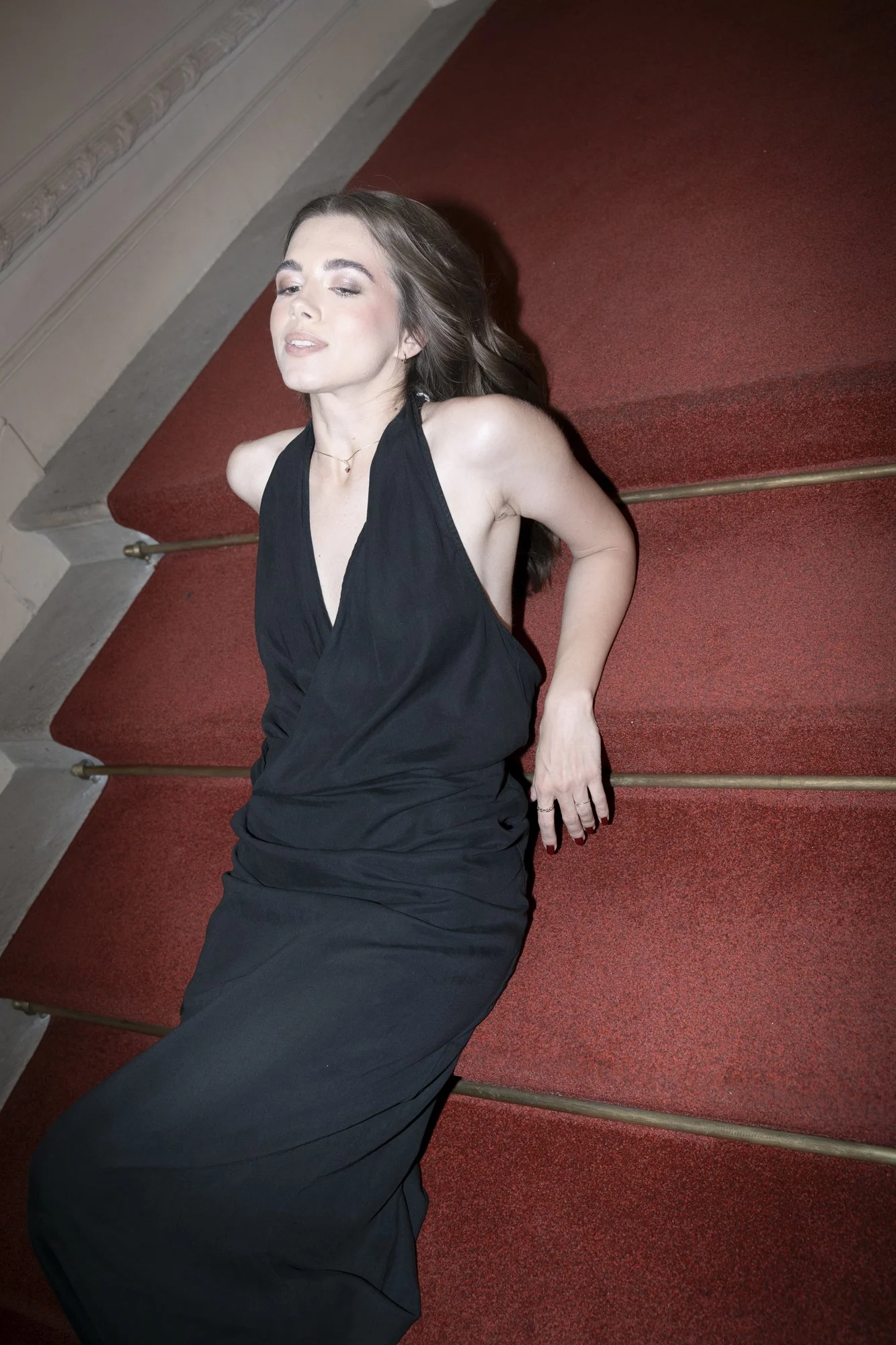 A woman in a black dress lying on a red carpeted staircase with gold handrails, eyes closed, and hair spread out.